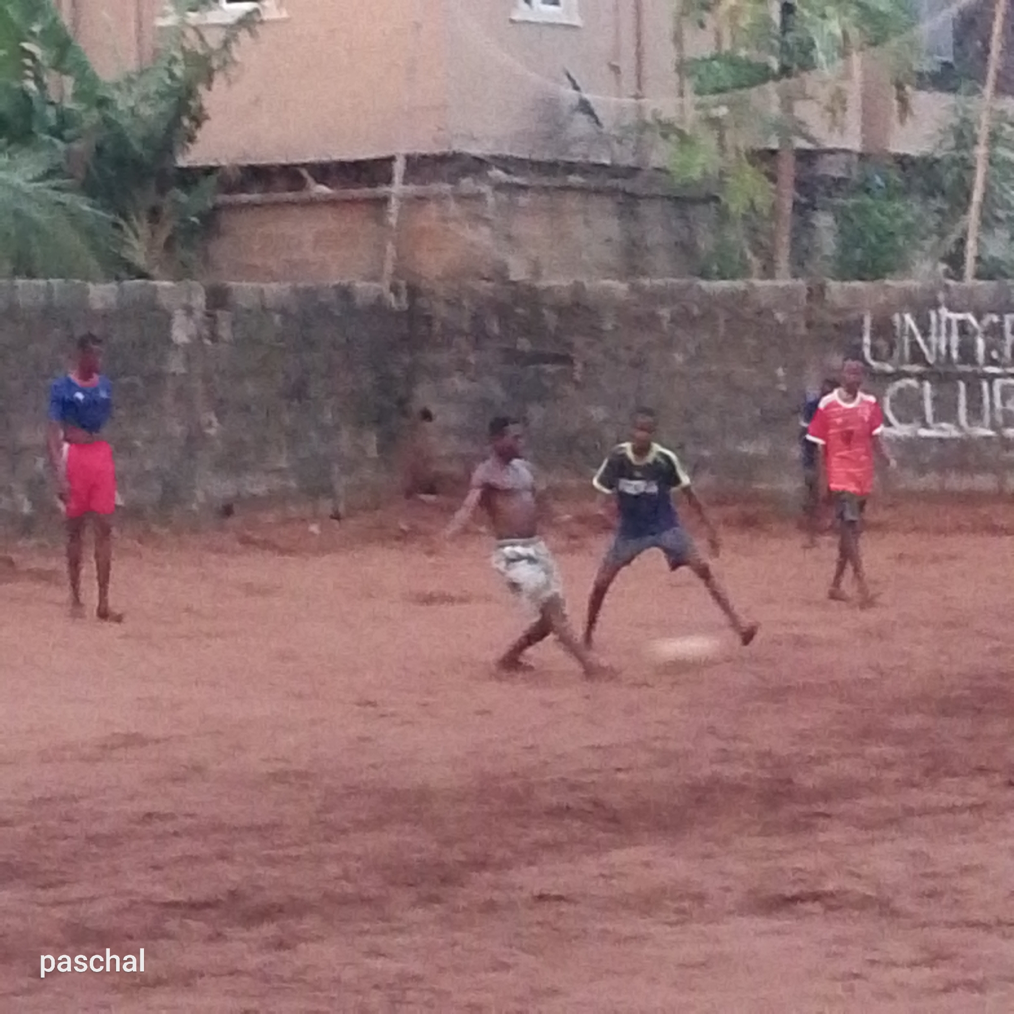 Four people are playing soccer on a dirt field. In the background is a weathered wall with the words "UNITY CLUB" painted on it. There are buildings and trees visible behind the wall. The photograph includes the name "paschal" in the lower left corner. Based on this information, the image is likely taken near a local Unity Club playing field. The location is possibly Ukwu-Abwa, Nigeria, as stated.