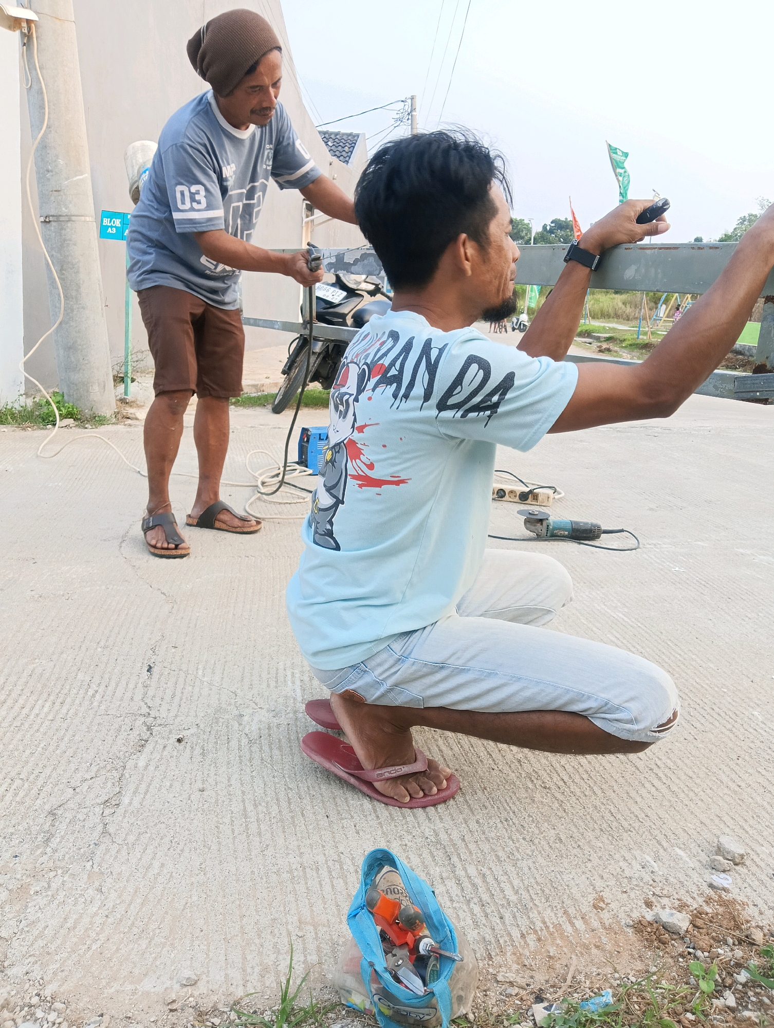 In Cikasungka, Indonesia, two men are working on a construction site. The man in the foreground, wearing a light blue t-shirt with a graphic design and light-colored pants, is crouching and holding a small black object, possibly a remote or a tool, in his right hand, while reaching out with his left hand to a metal railing. He is wearing red flip-flops. In the background, another man wearing a brown beanie, a grey t-shirt with "03" printed on it, and brown shorts, is standing and holding a tool. He is wearing brown sandals. A motorcycle is parked nearby. Tools, including an angle grinder, are laid out on the concrete ground. The environment appears to be an outdoor construction area with a cloudy sky overhead.