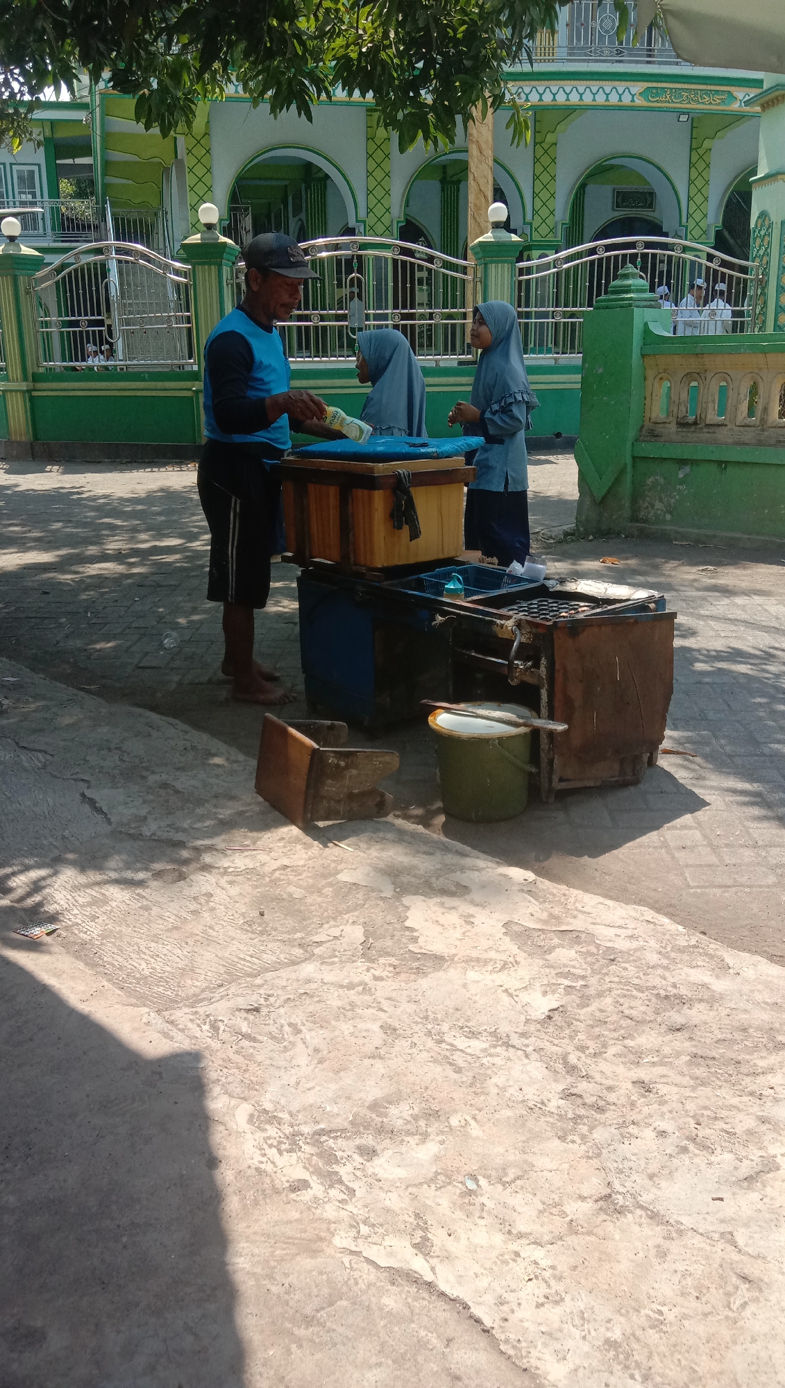 In Ngempit, Indonesia, under the shade of trees and in front of a mosque, a man wearing a blue long-sleeved shirt, black shorts with white stripes, and a dark baseball cap is handing money to a young person wearing a light blue hijab and matching tunic. Beside them, another young person in a similar light blue hijab and tunic stands, looking on. They are gathered around a wooden cart with multiple compartments. On top of the cart sits a yellow cooler. Below the cooler, a blue container is visible, and a rusty metal structure with what appears to be cooking apparatus is attached to the cart. To the left of the cart, a small wooden stool is overturned on the paved ground. A green bucket with a white lid is situated near the cart, and a shovel rests across it. The ground is paved with light-colored concrete. The mosque in the background features green and white architecture with arched windows.