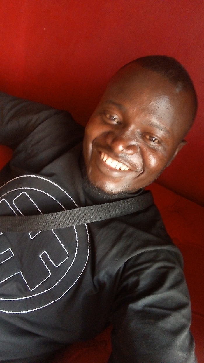 A smiling Black man in a black shirt with a white outlined graphic sits against a red backdrop. A black strap crosses his chest. The image is taken at an upward angle, showcasing his cheerful expression and attire. The focus is primarily on the man's face, highlighting his smile and the details of his features.