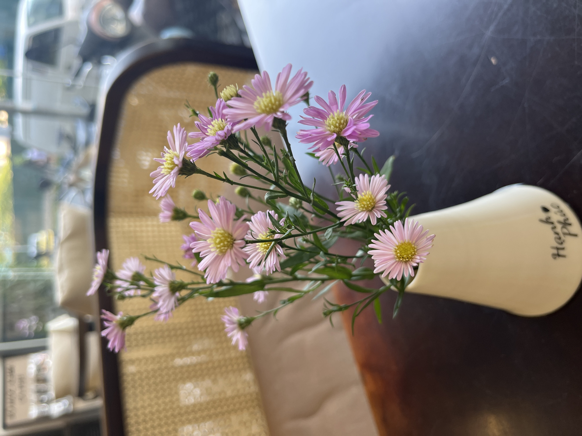 A close-up shot features a small, off-white vase holding a bouquet of pale pink daisies with yellow centers. The vase has handwritten script in Vietnamese. It is placed on a dark wooden surface, likely a tabletop. A chair with a woven wicker back is visible in the background, slightly blurred. Outside the window, a blurred cityscape suggests an urban setting, likely a café or similar establishment in Nha Trang Ward, Vietnam. The overall lighting is soft and natural.