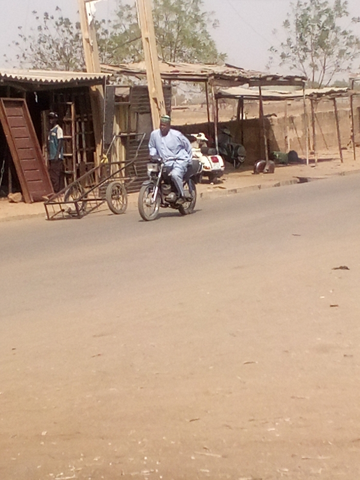 The image depicts a daytime street scene in Tudun Murtala, Nigeria. A male individual, dressed in a light blue traditional garment and a green cap, operates a dark-colored motorcycle moving from right to left across an asphalt road in the foreground. The road transitions to a dusty, unpaved shoulder on both sides.
On the left side of the road, a makeshift commercial stall with a corrugated metal roof stands open-fronted. Inside, a second male wearing a light blue and dark brown shirt is visible. Adjacent to the stall, a two-wheeled metal cart or trailer is positioned. Various metal objects, including what appears to be door frames, are stacked within and leaning against the stall structure. A long, brown, inclined object, possibly a ramp or large plank, rests against the stall's front.
On the right side of the road, further down the shoulder, another similar open-fronted shelter with a corrugated roof is present. A white scooter is parked on the dusty ground near this shelter, alongside other indistinct dark objects. A low, light-colored wall runs parallel to the road. The background reveals additional low-rise structures and sparse, light green foliage against a hazy sky. The overall environment appears dry and dusty.