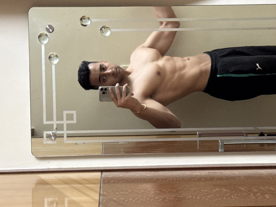 A man with dark hair takes a mirror selfie, showcasing his muscular physique. He has a toned torso and wears black athletic pants with a green stripe. The mirror has a decorative design with glass knobs and geometric patterns. The background features a wall and a wooden surface.