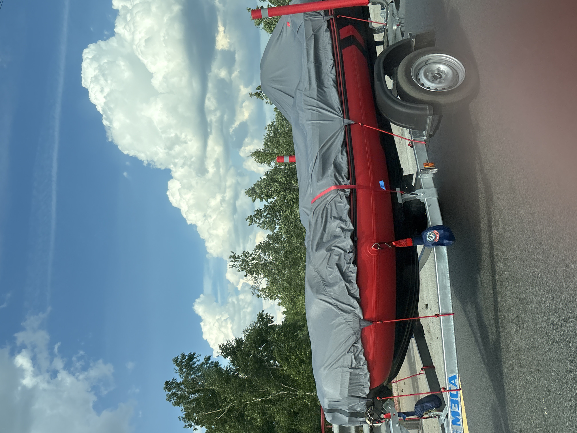 This image captures a red inflatable boat, covered by a grey tarp, positioned on a silver single-axle trailer, stationary on an asphalt surface in Razmetelevo, Russia. The boat's exposed hull is bright red, transitioning to a black lower section, and features a black stripe with a red chevron pattern along its side. The grey tarp, which custom-fits the boat, is secured by multiple red tensioning straps. The trailer exhibits the brand inscription "МЭЛА" (MELA) on its frame, a black wheel fender, and a silver wheel with a visible hub. Blue fabric securing elements are attached to the trailer near the front of the boat. The background consists of a dense arrangement of green deciduous trees under a clear blue sky, punctuated by scattered white cumulus clouds. Strong overhead sunlight is evident. A red horizontal structural element is partially visible on the far left, while two red cylindrical posts are positioned behind the boat on the right. No individuals or animals are present in the scene.