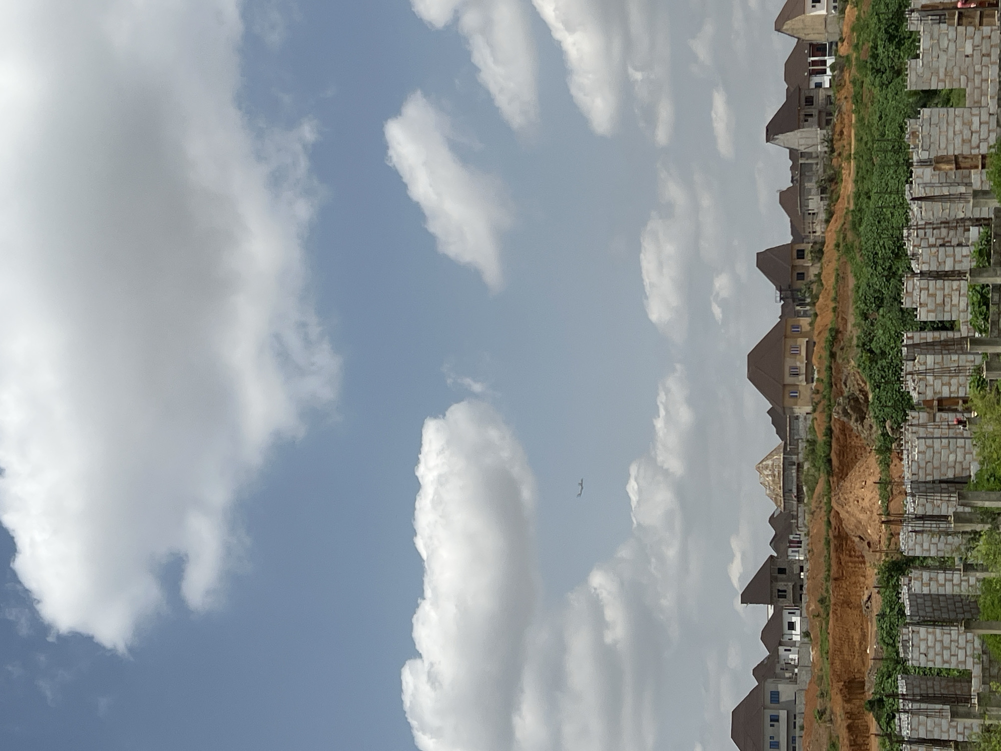 An aerial view captured from a distance shows a residential development under construction in Kubwa, Nigeria, beneath a partially cloudy sky. The foreground is dominated by the skeletal framework of unfinished buildings, characterized by concrete columns and cinder block walls. Beyond this construction zone, a landscaped hillside rises, dotted with completed and near-complete houses featuring tiled roofs and modern architectural designs. These homes are situated on tiered levels, with some appearing to be two-story structures. The landscape is a mixture of exposed reddish-brown earth and patches of green vegetation. In the sky, a commercial airplane is visible in the distance, appearing as a small, dark silhouette against the lighter clouds. The overall impression is one of ongoing urban expansion and development within a suburban setting.