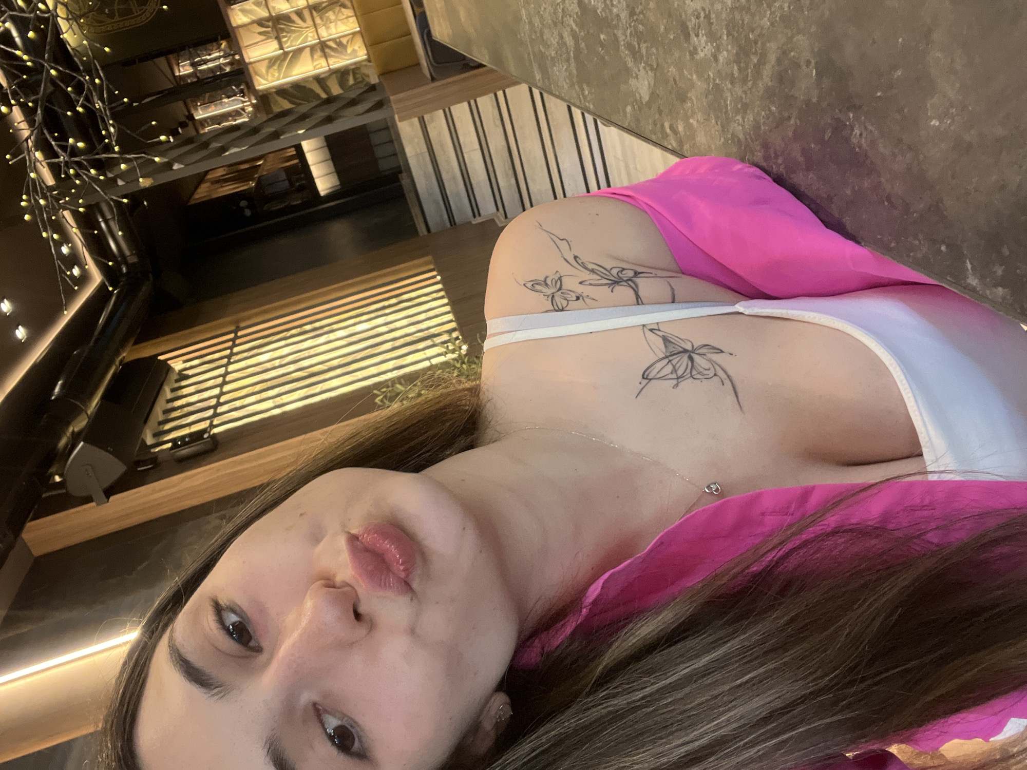 This is a vertical, close-up, selfie-style photograph taken indoors. The primary subject is a young woman with long, brown hair, her face turned towards the camera. She is wearing a bright pink garment draped over her shoulders and a white top. She has tattoos of butterflies on her upper chest and shoulder. Her lips are slightly parted, and she appears to be looking directly at the camera.
The background is out of focus but suggests an indoor setting with modern architecture. There are elements that look like interior lighting fixtures, possibly with yellow accents, and a textured wall or partition with horizontal lines. Stair railings are also visible in the upper portion of the image. The lighting in the scene is warm and ambient. The image seems to be taken during the day, given the brightness of the indoor lighting. The overall mood is casual and relaxed. No visible text is present in the image. The location context provided is Plovdiv, Bulgaria.