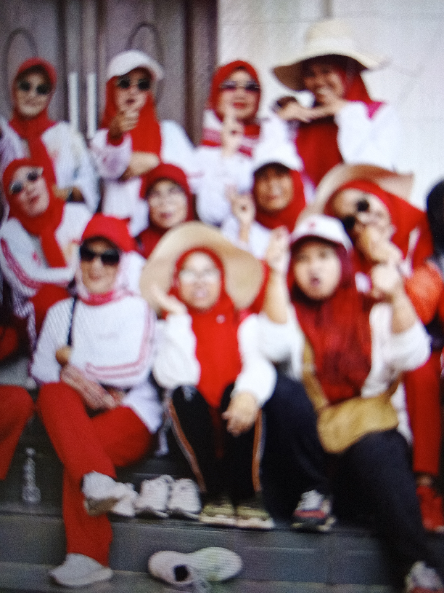 In Warungkiara, Indonesia, a group of individuals, mostly women, are gathered on steps. They are all wearing red and white clothing, with many sporting red headscarves or hats. Some are wearing sunglasses. The overall atmosphere appears to be festive or commemorative, possibly for a national event. The image is blurry, making it difficult to discern individual facial features or specific details of their attire. The environment is outdoors, with what appears to be a building or wall in the background.