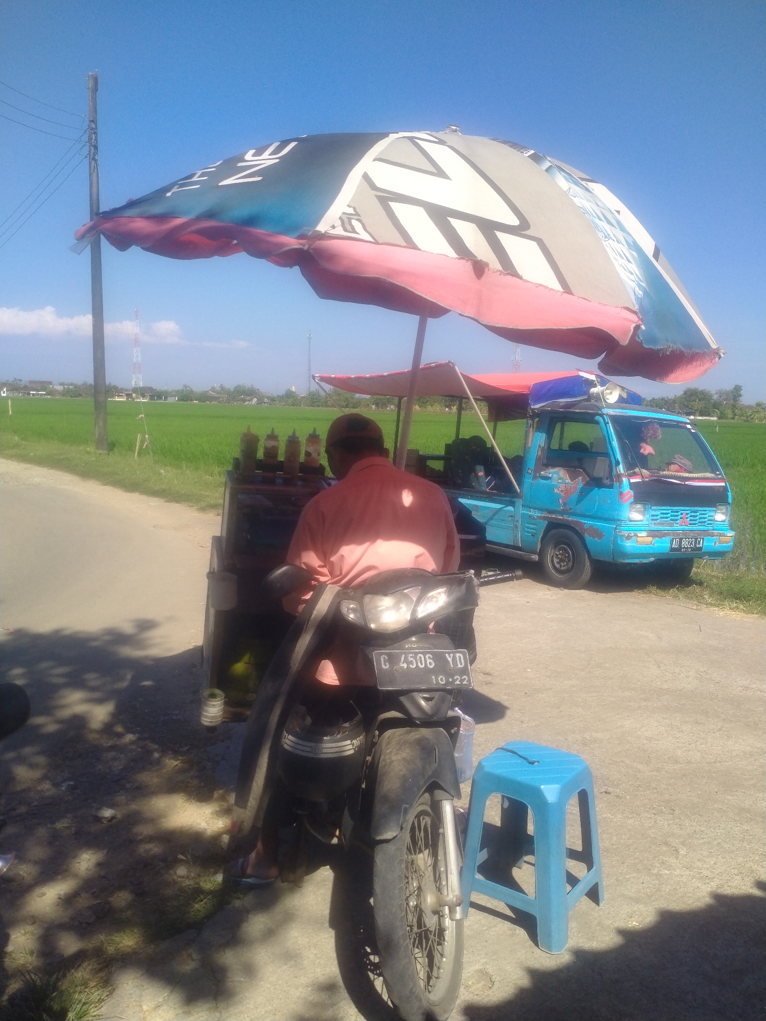 The image depicts an outdoor scene in the city of Begajah, Indonesia, likely on a roadside next to a rice field. The focus is on a food cart set up with a motorcycle and a small blue truck. A man wearing a light orange shirt is seen from behind, standing at the food cart, which is laden with bottles of condiments. The food cart is attached to a motorcycle, the rear of which is partially visible. To the right of the cart, a bright blue plastic stool is placed on the ground. A light blue Mitsubishi truck is parked behind the cart, its front visible. The truck has a license plate that reads "AD 8823 CA". A large umbrella, predominantly white with black and pink accents, is set up over the food cart. In the background, green rice fields stretch towards the horizon under a clear blue sky. A utility pole with electrical wires stands on the left side of the image.