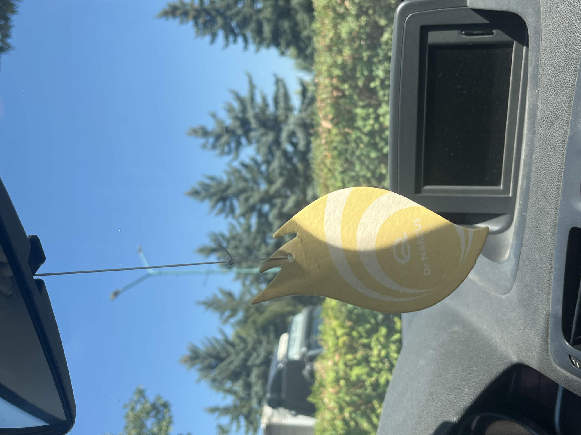 An interior view from a vehicle's dashboard looking through the windshield. The scene is illuminated by bright sunlight under a clear blue sky. A yellow, flame-shaped air freshener, bearing the brand "Dr. Marcus" and a stylized circular logo, hangs from a thin string in the foreground, obscuring a portion of the view. The air freshener has concentric yellow lines on its surface. Partially visible on the dashboard is a dark, rectangular display screen. In the middle ground, a line of green bushes and trees is visible. Beyond the foliage, several evergreen trees stand tall, and in the distance, a construction crane is visible against the sky, suggesting an urban or suburban environment. The background also shows the rear of vehicles, indicating a road or parking area. The specific location of Targovishte, Bulgaria, is not identifiable from the visual elements present in the image.