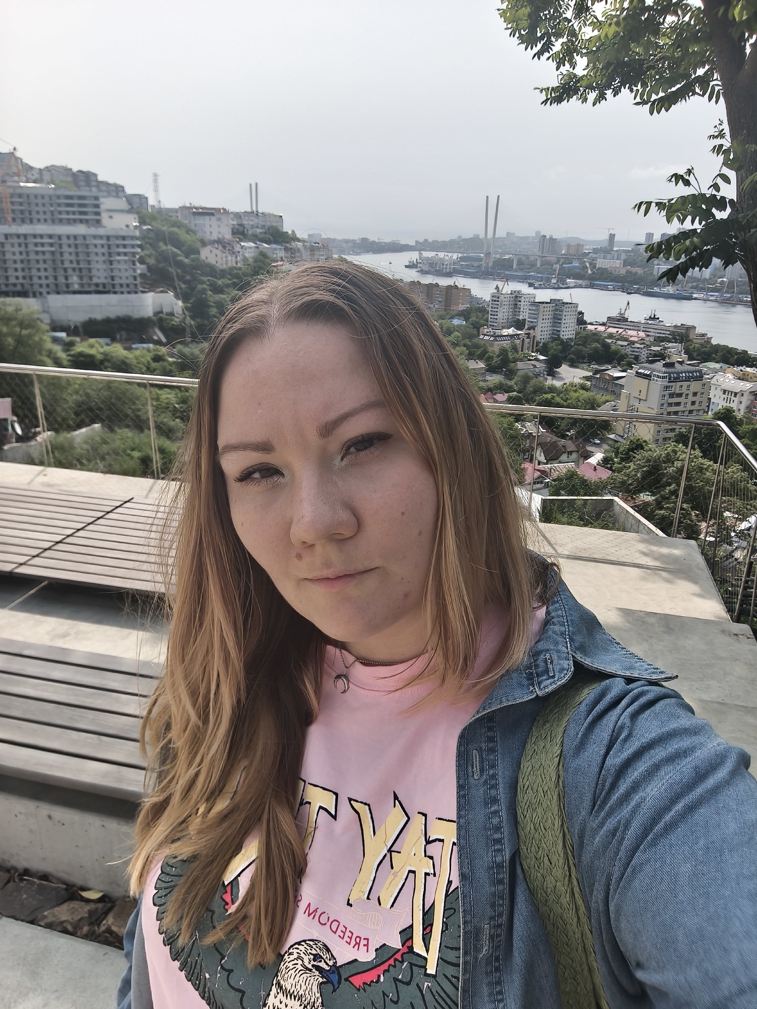 A woman with blonde hair is taking a selfie with a scenic background of Vladivostok, Russia. She is wearing a pink t-shirt with a graphic of an eagle and a denim jacket. A green backpack is visible. The background shows buildings, a body of water, and bridges. The setting appears to be an elevated vantage point overlooking the city.