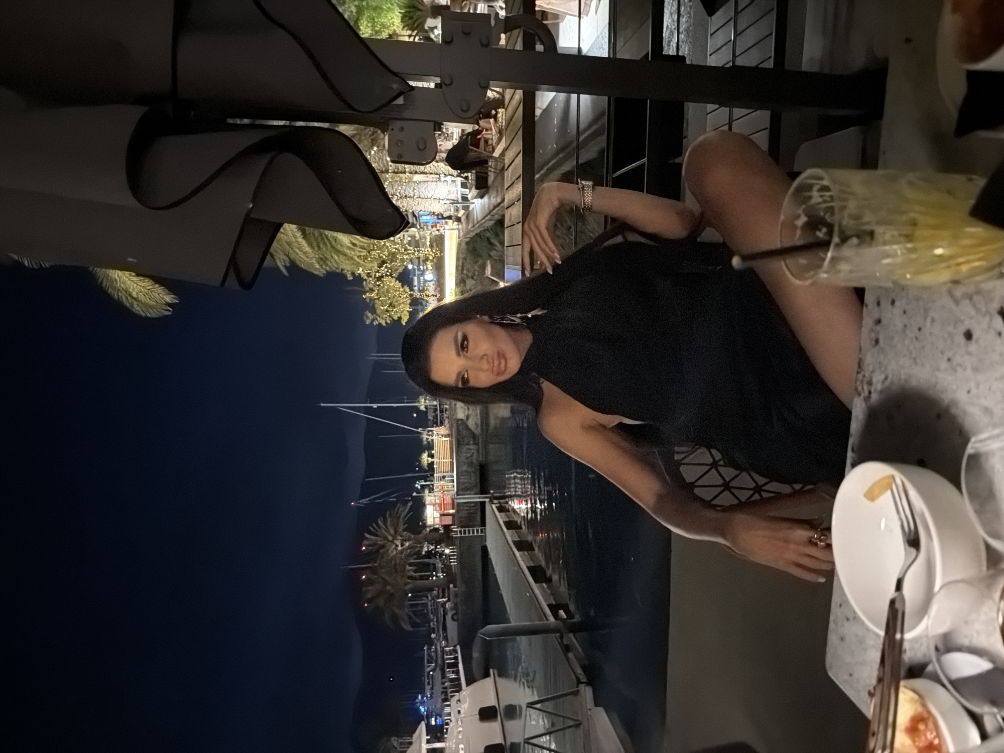 A woman in a black dress is seated at a table outdoors at night, with a marina in the background. The woman has long dark hair, is wearing makeup, and jewelry, including earrings and bracelets. She is looking to her left with her legs crossed. On the table in front of her are bowls of food, a fork with a piece of food, and a glass with a straw. The marina is lit with lights and features boats and palm trees. The location is Tivat, Montenegro.