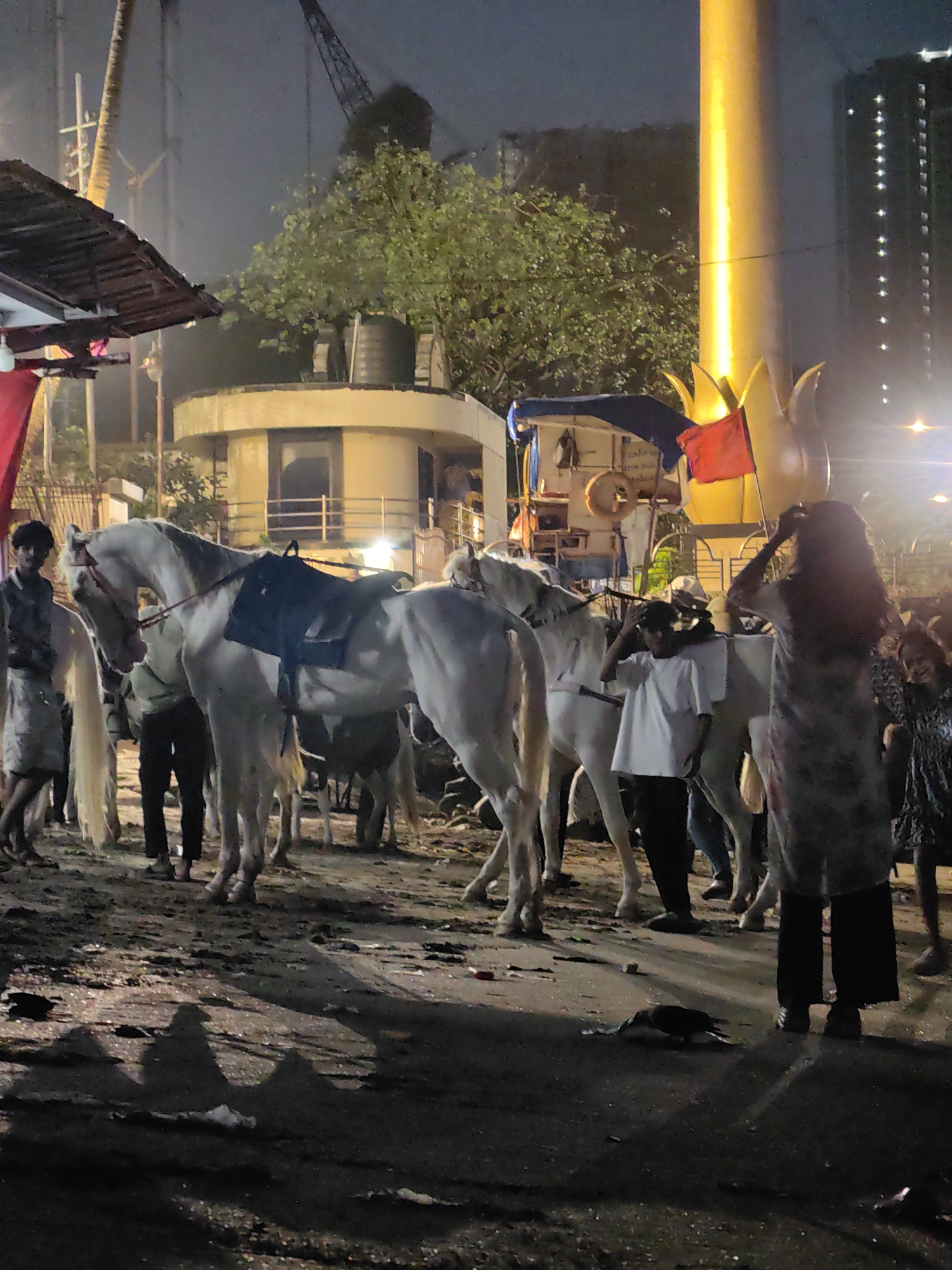 The image depicts a nighttime scene in a Mumbai, India, setting, likely near a street or event area. In the foreground, several white horses are present, some equipped with saddles. A group of people, both male and female, are visible interacting with the horses and each other. One man, wearing a light-colored shirt and dark pants, stands near a horse. Another person, with long dark hair and wearing a patterned dress, is seen from behind, with their hands near their head. A young person in a white t-shirt and dark pants is also visible.
In the background, there is a large, illuminated cylindrical structure that appears to be a light pole or part of a monument, casting a bright yellow glow. Behind that, a tall building with illuminated windows can be seen. Trees and foliage are present, adding to the urban greenery. Structures resembling a canopy or shelter with a red cloth are also part of the scene. A crane is visible in the distance, suggesting ongoing construction or industrial activity. The ground appears to be a dirt or paved surface with some debris. A bird is seen scavenging on the ground in the lower right portion of the image. The overall atmosphere suggests a lively, possibly festive, environment at night.