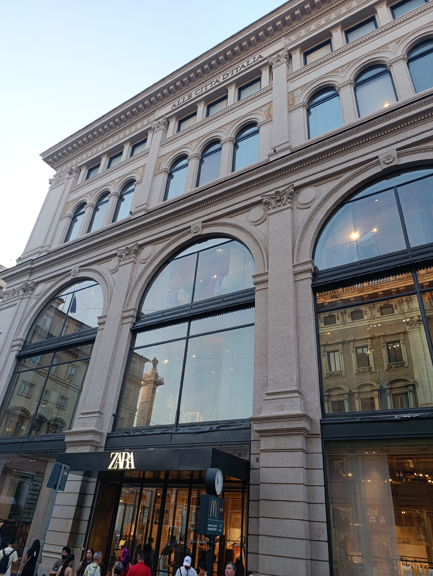 This image captures the facade of a Zara store located in the city of Rome, Italy. The building, characterized by its neoclassical architecture, features multiple levels of arched windows. The ground level boasts large, expansive glass windows that reflect the surrounding cityscape and interior of the store. A prominent "ZARA" sign is illuminated above the entrance. A McDonald's sign is also visible nearby. Several people are seen walking on the sidewalk in front of the store. The sky above is clear, indicating daylight.