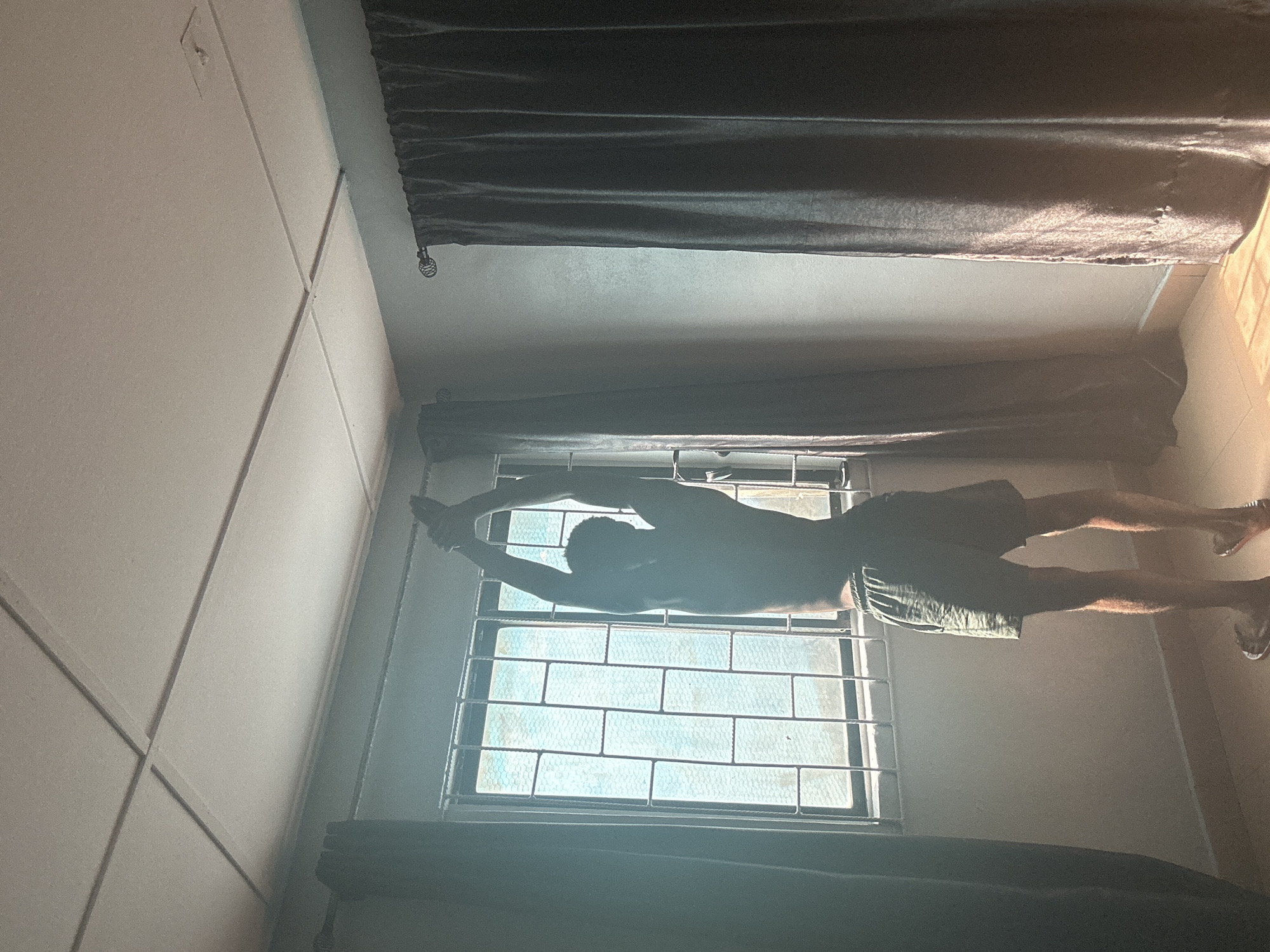 A person is standing in a room with their arms raised above their head, in front of a window. The window has metal bars across it. The room has dark curtains on either side of the window and a tiled ceiling. Sunlight is coming through the window, casting shadows on the floor. The person is wearing shorts. The scene appears to be located in Badagry, Nigeria.