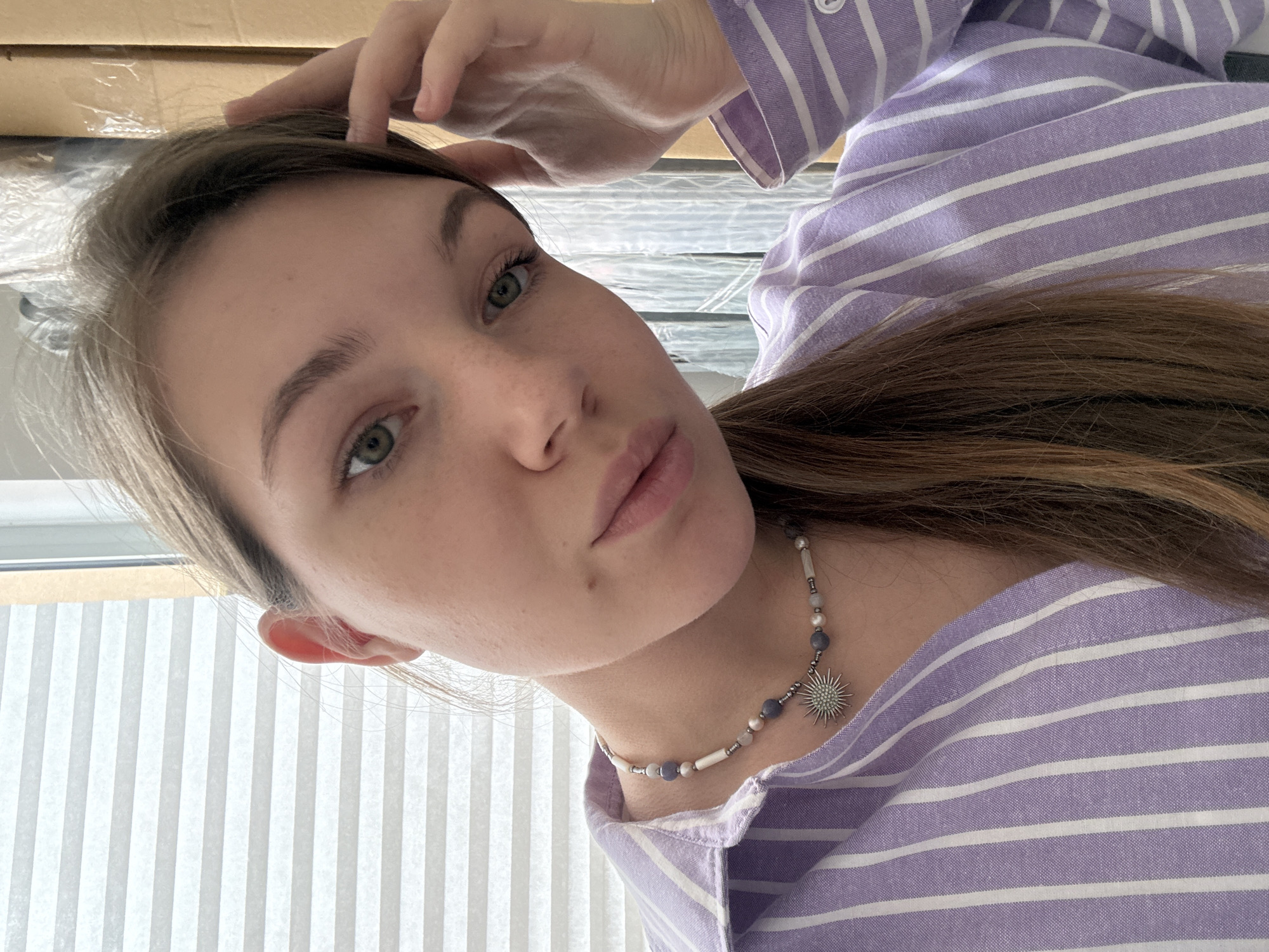 A young woman with fair skin and light brown hair is pictured in a close-up selfie. She is wearing a light purple and white striped shirt and a beaded necklace with a sun-shaped pendant. Her left hand is touching her forehead. The background includes a window with blinds and what appears to be a box. The image does not provide information related to the city of Vracar Urban Municipality, Serbia.