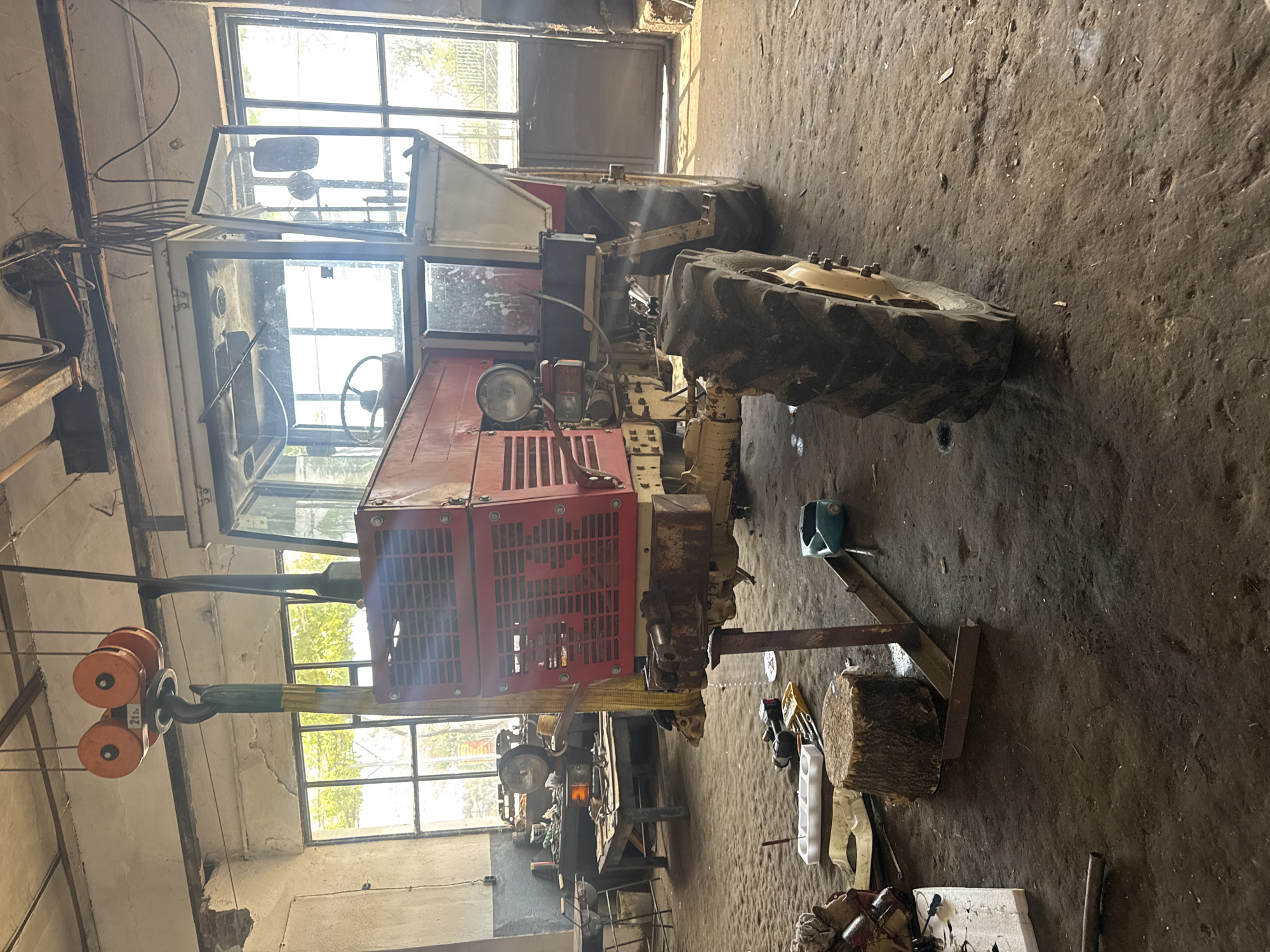 The image displays a red and white tractor undergoing maintenance or repair inside an industrial or agricultural workshop in Sarnevo, Bulgaria. The tractor is horizontally oriented in the frame, with its front section elevated and resting on a robust metal stand. A hoist system, featuring orange pulleys and a yellow strap, is attached to the front of the tractor. The hoist clearly displays the text "2t," indicating a 2-ton lifting capacity.
The tractor appears to be an older model, showing signs of significant use and wear, including faded paint and some rust. It features a red engine cover with a prominent grille, a single front headlight, and a cabin enclosed by multiple glass panes. A steering wheel is visible within the cabin. The vehicle has large, deeply treaded rear tires and smaller front tires.
The workshop environment is practical and well-used. The floor is a rough concrete or dirt surface, visibly stained and worn. Abundant natural light enters the space through several large, grimy multi-pane windows located along the walls, indicating it is daytime. In the background and around the tractor, various tools and workshop items are scattered, including a wooden log, plastic containers, and other indistinct equipment. Cables are visible along the walls, reinforcing the industrial nature of the setting. No people are present in the scene. The overall impression is that of an active repair facility.