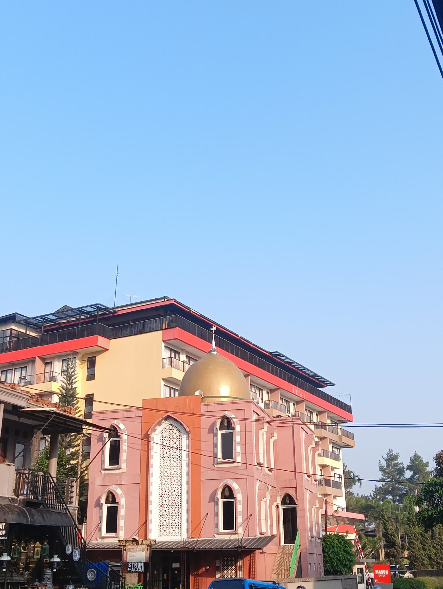 In Cisarua, Indonesia, a pink mosque with a gold dome stands adjacent to a multi-story building. The mosque features arched windows and decorative white latticework on its facade. Above the mosque, the multi-story building is painted in shades of yellow and red and has balconies and a rooftop structure with a glass canopy. Power lines are visible crossing the frame, and trees are present in the background. A sign in Indonesian that reads "NASIGORENG BANDUNG" is visible on the lower part of the mosque.