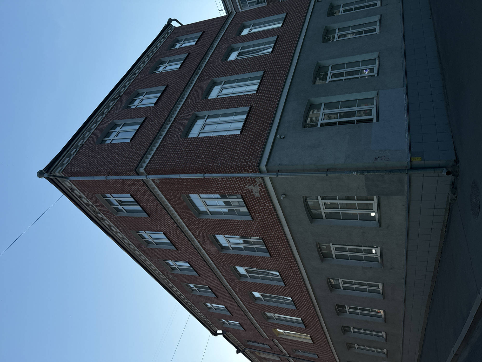 The image shows a red brick building with a light blue lower story, viewed from a street corner at dusk. The building has multiple floors and many windows. The sky is clear and light blue. There are no people visible in the image. The building appears to be in a residential or commercial area, with a street and sidewalk in the foreground. The architecture suggests a post-Soviet or pre-revolutionary style. The building is located at Bolshoy Trekhsvyatitelsky Lane, 2/1, in Moscow, Russia.