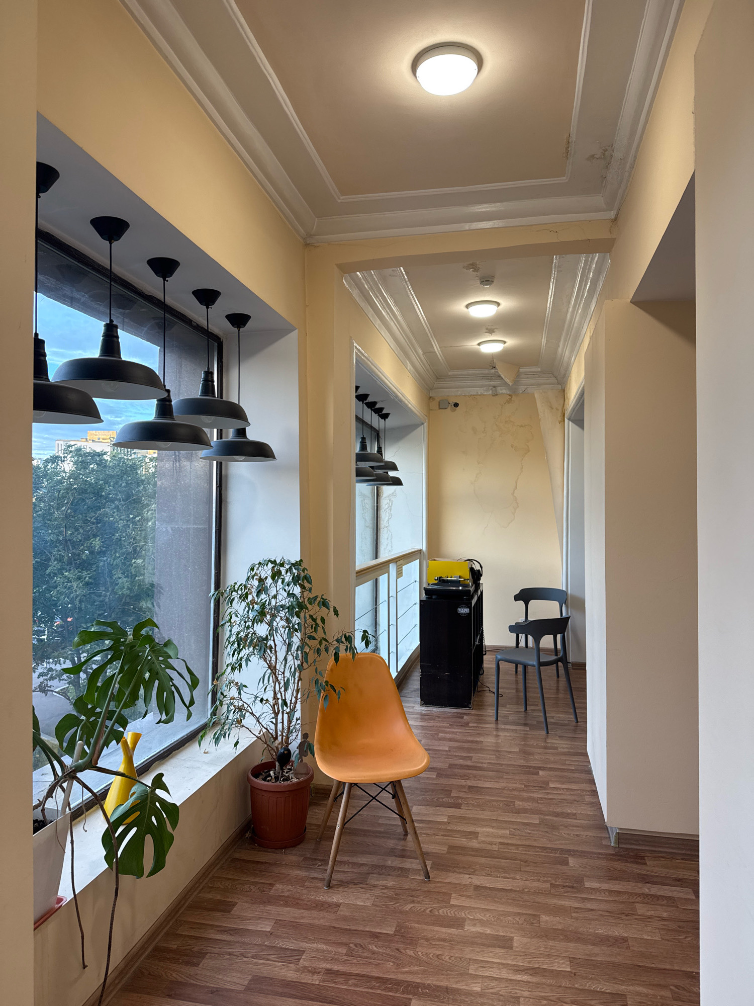 The image depicts a hallway with a large window on the left. The hallway is illuminated by several ceiling lights and a row of pendant lights hanging near the window. The walls are painted a pale yellow, and the ceiling has decorative molding and some water damage. The floor is covered with wood-look laminate flooring.
On the windowsill, there are several potted plants, including a large Monstera deliciosa and a Ficus tree. A bright yellow vase is also visible. An orange molded plastic chair with wooden legs is placed in the foreground, facing the window.
Further down the hallway, a black cabinet with what appears to be audio equipment, including a turntable and headphones, is situated. Two dark gray chairs are placed behind the cabinet. The hallway leads to an open doorway at the far end.
The view through the window shows trees and buildings, suggesting an urban environment. The architecture of the buildings outside, combined with the style of the interior molding, could be consistent with older buildings found in Moscow, Russia. The overall impression is of an indoor space that is perhaps a cafe, studio, or office hallway, with a blend of modern furnishings and some signs of wear and tear.