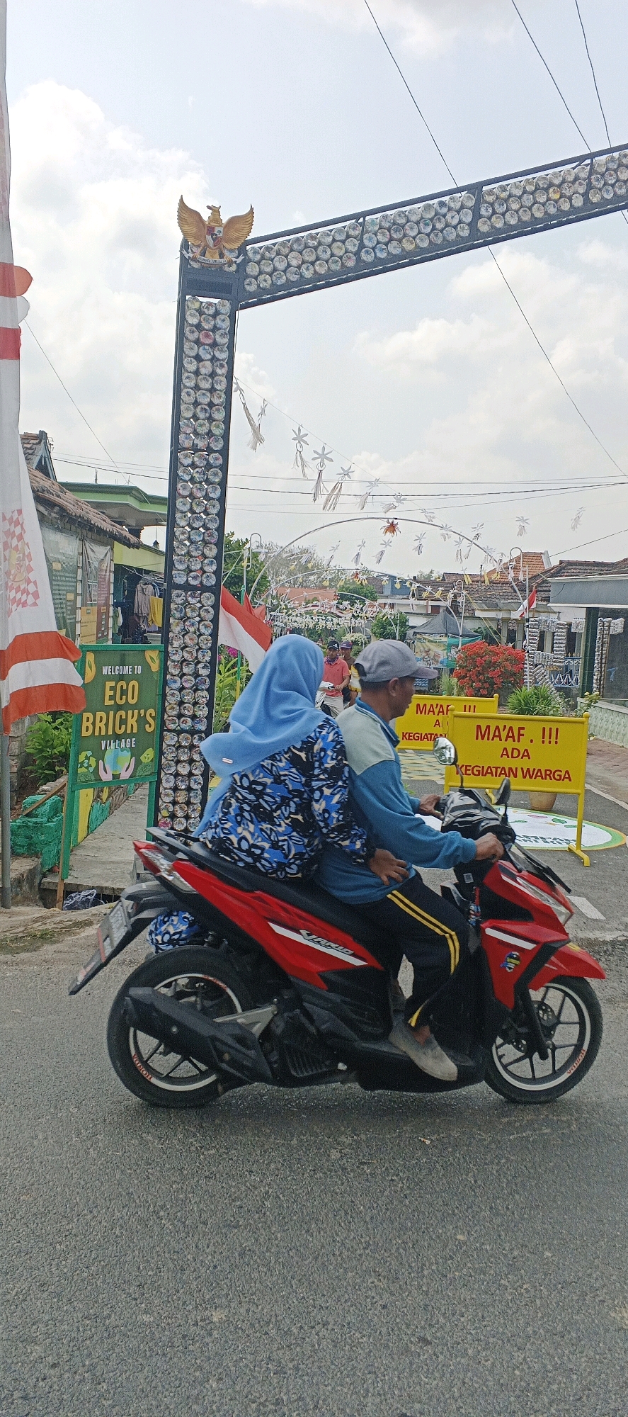 In the city of Bluri, Indonesia, two individuals are riding a red motorcycle on a paved road. The driver, a man wearing a grey baseball cap and a blue and grey long-sleeved shirt with yellow stripes on the pants, is operating the vehicle. The passenger, a woman wearing a blue hijab and a blue and black patterned jacket, is seated behind him. The motorcycle is moving past a decorated archway made of what appear to be plastic bottles. To the left of the archway, a green sign reads "WELCOME TO ECO BRICK'S VILLAGE". Behind the archway, a yellow banner with red lettering states "MA'AF. !!! ADA KEGIATAN WARGA" which translates to "SORRY. !!! THERE ARE COMMUNITY ACTIVITIES". The environment includes buildings with red-tiled roofs, trees with red flowers, and overhead power lines. The sky is cloudy.