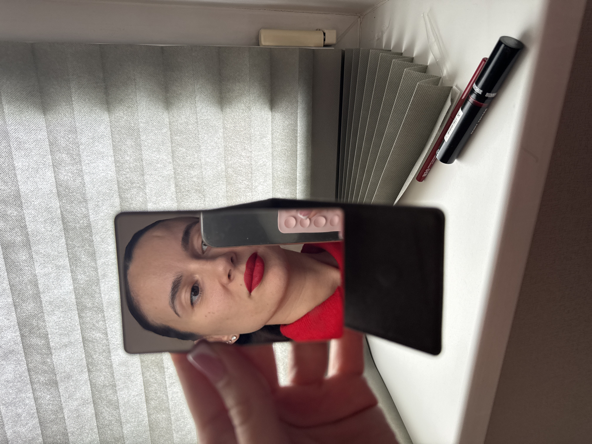 A photograph taken indoors in what appears to be Chaikovsky, Russia, shows a woman's face reflected in a small handheld mirror. The mirror is held in a hand with visible fingernails, reflecting the woman's face, adorned with red lipstick and a red top. The background shows a window with closed white blinds. Adjacent to the window sill are cosmetic items – a mascara and a lip liner. The composition suggests a personal grooming or beauty routine is taking place.