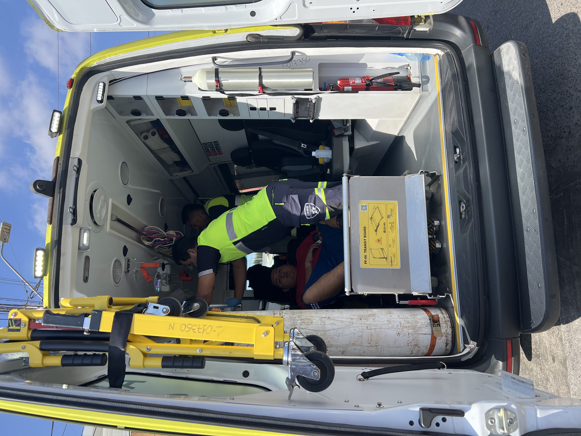 The image shows the interior of an ambulance, likely in Arica, Chile, given the presence of "SAMU" (Servicio de Atención Médica de Urgencia, a Chilean emergency medical service). An individual, presumably a patient, is lying on a stretcher or transit board inside the ambulance. A medical professional wearing a high-visibility yellow and black uniform is attending to the patient. Medical equipment, including an oxygen cylinder and a fire extinguisher, are visible within the ambulance. The stretcher has a serial number on it 1-017350 N. The stretcher platform is labelled PF-06 TRANSIT BOARD and has an image of the folding stretcher with a max load of 220 kg.