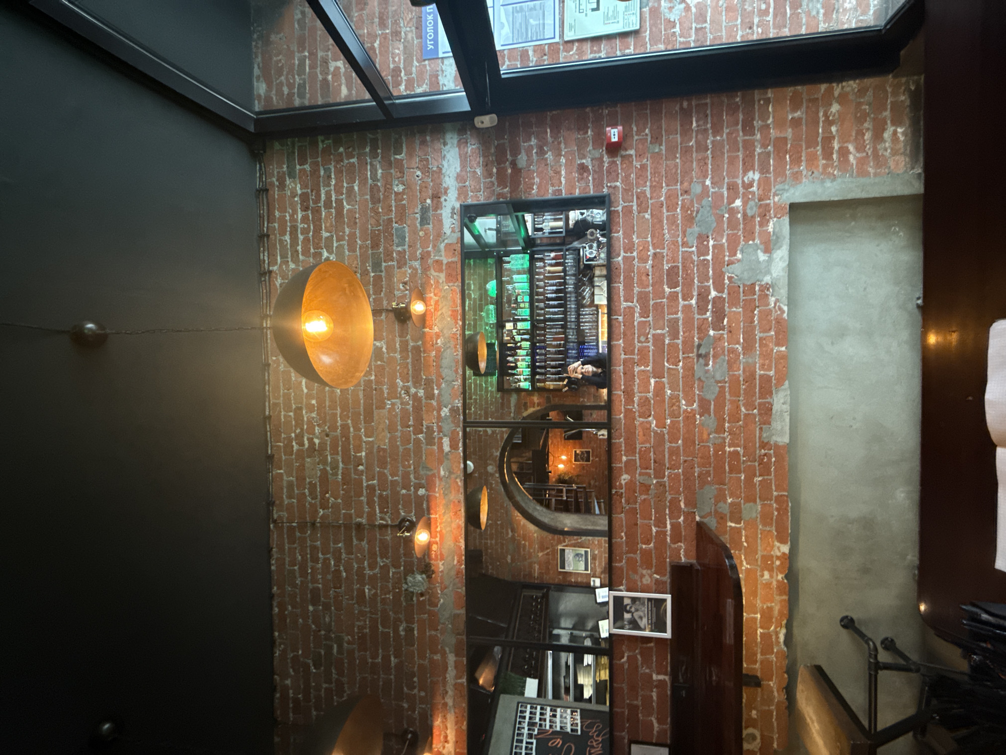The image captures an interior scene, possibly within a restaurant or bar. A dominant feature is a textured brick wall with a vintage aesthetic, exhibiting a mix of red and grey hues. Affixed to the wall is an elongated mirror that offers a glimpse into another area, reflecting what appears to be a wine display, furniture, and other decor elements.
On the left side of the brick wall, there's a dark grey or black painted wall with a spherical lamp extending from it. The presence of metal framing indicates a possible glass or skylight ceiling structure overhead.
A doorway is partially visible on the right side of the brick wall. Near the bottom center, a shelf displays framed photos and various items. The lighting is warm and uneven, creating a cozy yet somewhat dimly lit atmosphere. The location is potentially in Moscow, Russia, but the image alone doesn't provide explicit location confirmation.