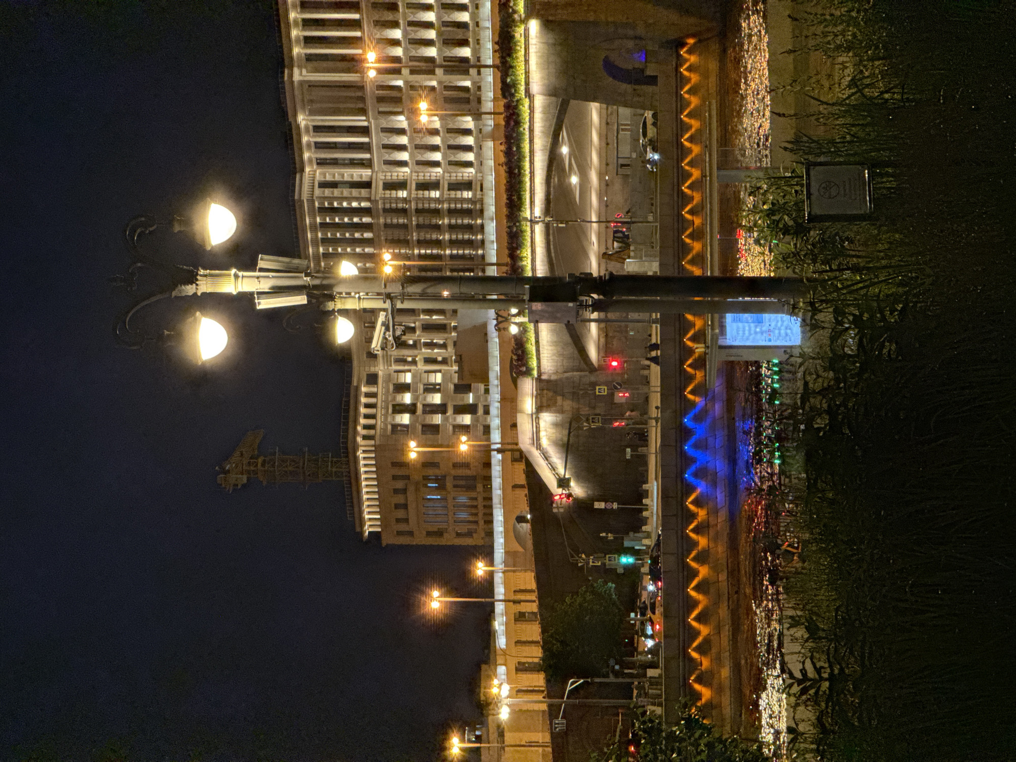 The image depicts a nighttime scene in Moscow, Russia, likely along the Moskva River, near a bridge. In the foreground, lush dark vegetation obscures the lower part of the frame. The water is illuminated by an array of lights: an orange, zigzagging pattern runs along the embankment, and further out, scattered lights in shades of blue and green reflect on the surface, suggesting a fountain or decorative lighting installation.
Dominating the midground is a bridge with a distinct architectural style, featuring illuminated arches and a walkway lined with flowerbeds. Traffic lights are visible, with one showing red and another green, indicating ongoing vehicle movement on the bridge or approaches. Several cars are present, some with headlights on, traversing the bridge.
A prominent street lamp with multiple globes stands in the mid-right of the frame, casting a warm glow. Behind the bridge and street lamp, a large, classical-style building with numerous illuminated windows and columns is visible. To the left of this building, an older, multi-story structure with a patterned facade is also lit up. In the distance, a construction crane with a yellow tower is silhouetted against the dark night sky.
The overall atmosphere is urban and vibrant, showcasing the city's illuminated architecture and waterfront at night. No specific individuals are clearly identifiable or engaged in notable actions. The scene captures the architectural grandeur of Moscow juxtaposed with the dynamic lighting of its urban infrastructure.