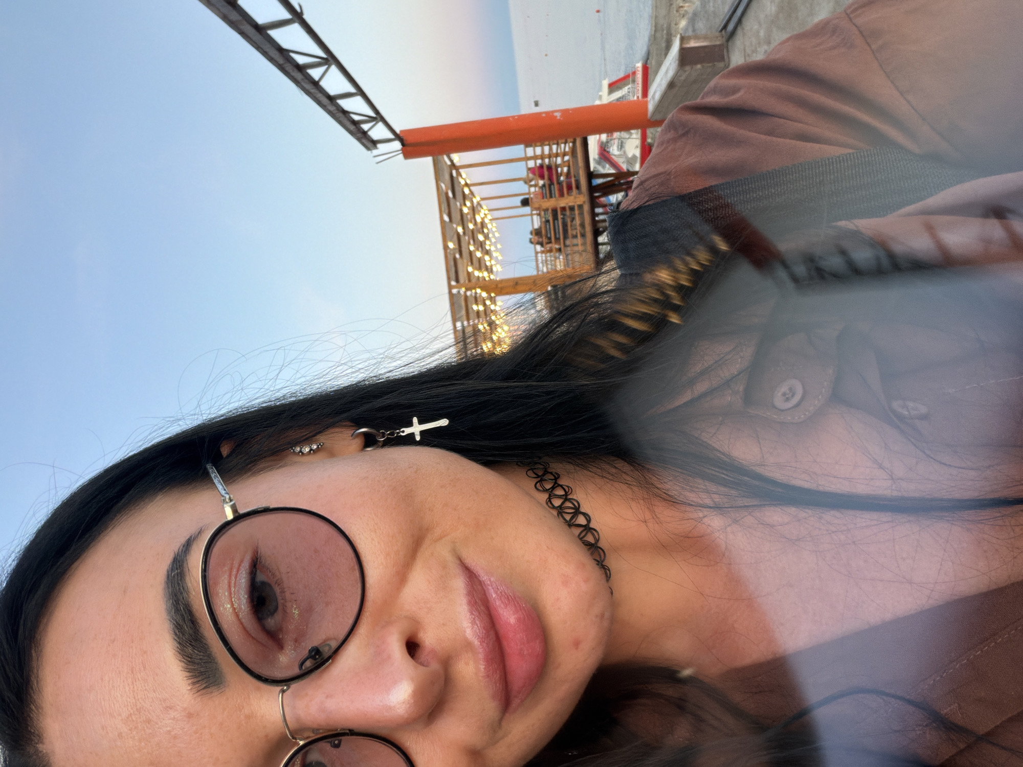A medium shot captures a young woman with long, dark hair and round, rose-tinted glasses. She is wearing a brown shirt and a black choker necklace. Her left ear is adorned with a cross-shaped earring. She is smiling at the camera. In the background, a wooden structure with string lights, several people, and the sea are visible. The sky is clear and pale blue. The scene appears to be an outdoor dining area by the water.