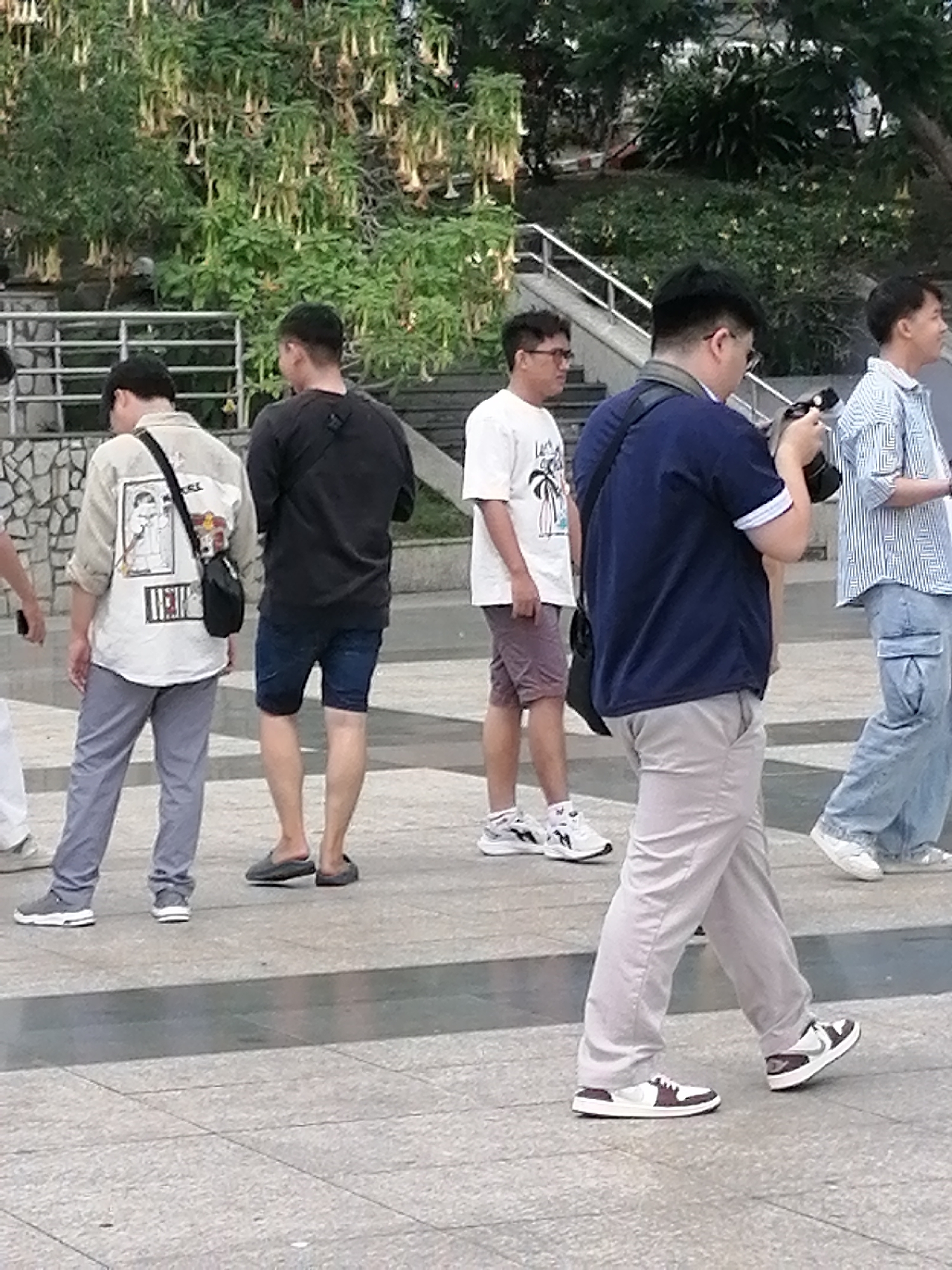 The image captures a group of people outdoors, likely in an urban setting with lush greenery in the background. In the foreground, four individuals are visible from the waist down, walking on a paved surface. The person on the far left is wearing grey trousers and white sneakers. Next to them is another person wearing blue shorts and dark slip-on shoes. The central figure is wearing light-colored trousers and two-toned sneakers. To the right, a person is wearing blue cargo pants and white sneakers. The background features a stone wall, a metal railing, and trees with hanging flowers, possibly in a park or garden area. The overall impression is that of a casual outing in a city, potentially Da Lat, Vietnam, known for its floral landscapes and mountainous terrain.