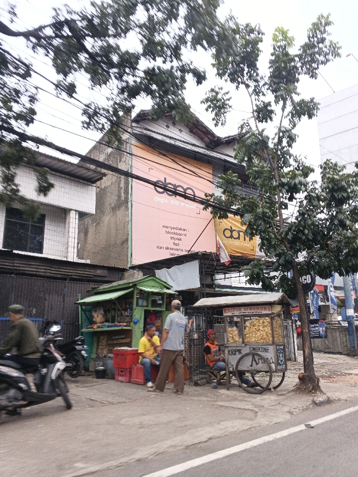 The scene depicts an urban street environment in Bandung City, Indonesia, under an overcast sky. An asphalt road occupies the foreground, bordered by a concrete sidewalk on the right. Numerous overhead power lines traverse the upper portion of the frame.
On the left, a two-story building with white tiled cladding is partially visible. Centrally located in the background is a multi-story building featuring a large, light-pink banner displaying "dema depo alat mebel" (dema furniture tools depot) and listing "blockboard, triplek, tipis, dan aksesoris mebel lain-lain" (provides: blockboard, plywood, thin, and other furniture accessories). A smaller yellow banner with similar text is below it. This building has a traditional sloped roof. To its right, a modern building is partially visible.
On the sidewalk, a green-painted vendor kiosk is positioned. Adjacent to it are stacked red plastic crates. To the right of the kiosk, a two-wheeled wooden cart, labeled "KRIPIK SINGKONG" (Cassava Chips) and "Aroma," displays bags of chips. A white tarp is strung above this vending area. A small tree with exposed roots stands next to the chip cart.
Four male individuals are present. On the far left, a person in a green top and cap operates a dark-colored scooter, moving leftward and exhibiting motion blur. Near the green kiosk, one male subject in a yellow t-shirt and blue jeans is seated on a red crate, facing right. Another male subject, wearing a light blue t-shirt and brown pants, stands with his back to the camera, appearing to engage the seated individual. Near the chip cart, a third male subject in an orange t-shirt and dark pants is seated on a low stool, facing left.