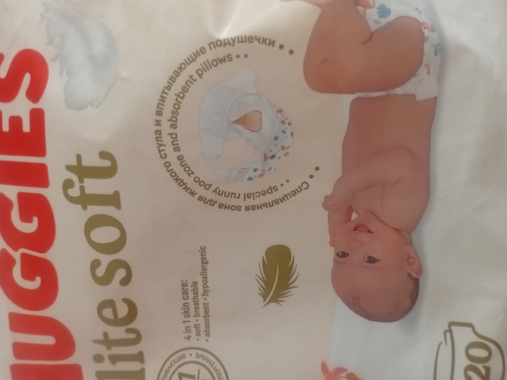 The image is a close-up shot of a package of Huggies Elite Soft diapers, featuring a sleeping baby on the lower portion of the package. The branding and product name are prominently displayed at the top. Text on the package describes the product's features, such as "4 in 1 skin care: soft, breathable, absorbent, hypoallergenic." There is also text in Russian and English that translates to "special runny poo zone and absorbent pillows." The background is a soft, neutral color, and the overall impression is one of cleanliness and comfort. There is no information or visual cue within the image to suggest a specific location in the city of Малая Владимировка ауылдық округі, Kazakhstan.