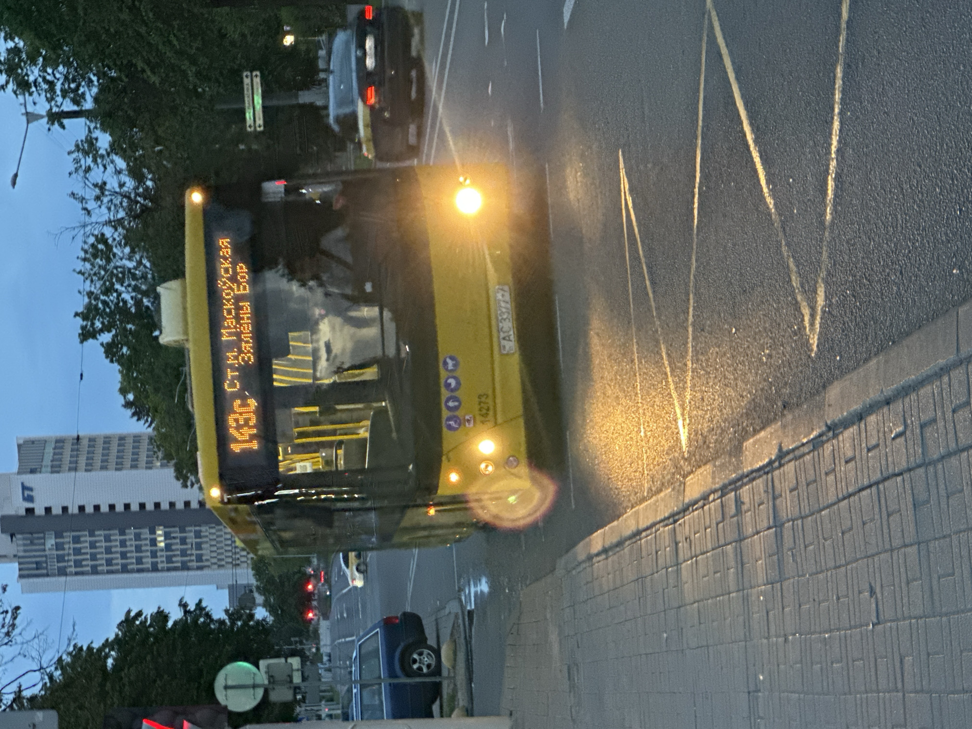 This image captures a yellow public transit bus, identified as fleet number 14273, displaying route 143c with the destination "ст.м. Маскоўская Зялёны Бор" (Maskovskaya Metro Station - Zialiony Bor). The bus is equipped with a Belarusian yellow license plate, AC 3377-7, and features international accessibility pictograms on its front. Its headlights are illuminated, casting a warm glow on the dark asphalt road, which features white lane markings and yellow zig-zag lines typical of a bus stop.
The bus is positioned on the right side of a multi-lane road, adjacent to a grey paved sidewalk. In the background, a tall, modern white skyscraper with a distinctive facade and a visible "БТ" (BT) logo on its upper section stands prominently. This building is identifiable as the **Belarusbank Headquarters at Dzerzhinsky Avenue 18, Minsk, Belarus**, despite the logo appearing as "БТ" rather than "ББ" or "БЕЛАРУСБАНК" due to perspective or lighting. The bus is located on Dzerzhinsky Avenue, in the vicinity of the Maskovskaya Metro Station.
To the left, a dark blue station wagon is parked near the sidewalk. Further down the road, a white car and a dark sedan with illuminated red taillights are visible. A traffic light with a red signal is visible on the left, along with a green pedestrian signal. The sky is a twilight blue-grey, indicating either dusk or early morning. Lush green trees line the street on both sides. A single person, likely the driver, is discernible behind the bus windshield.