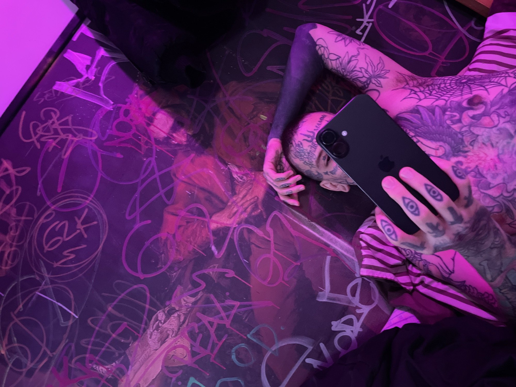 A person lies on a surface covered with graffiti, holding a black smartphone. The person is heavily tattooed, including their face, neck, torso, arms, and hands. The tattoos include designs like eyes, flowers, and spiderwebs. The lighting is primarily pink and purple. The scene appears to be indoors. There is no specific information available that would place the image definitively in Buenos Aires, Argentina.