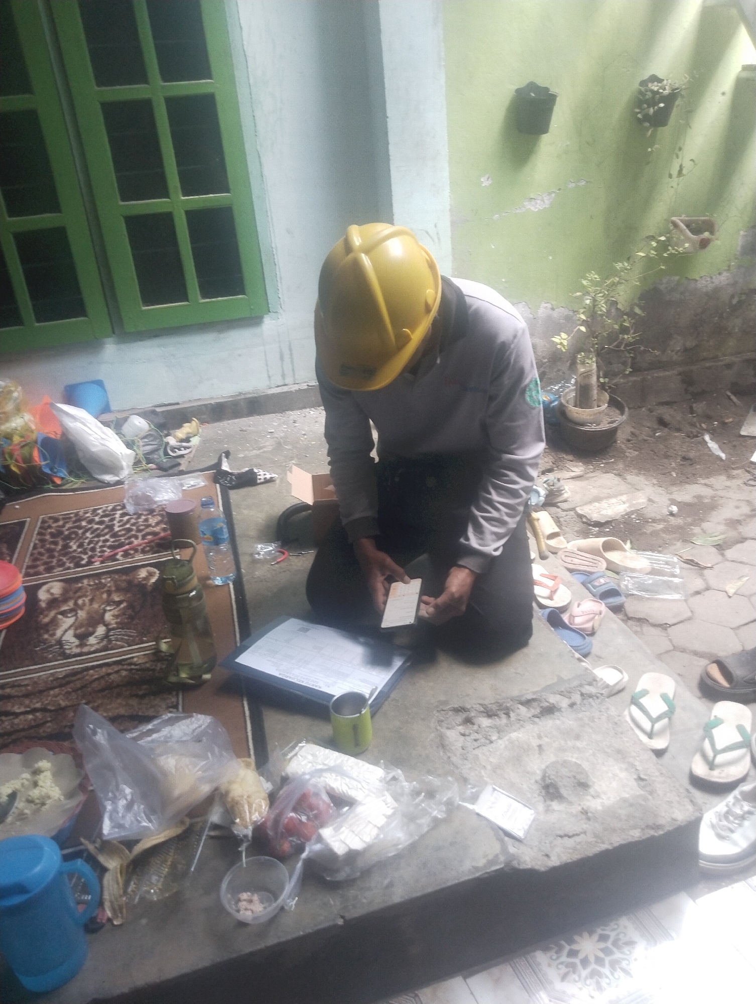 The image depicts an outdoor scene in what appears to be a courtyard or alleyway in Wanasaba Daya, Indonesia. A person wearing a yellow hard hat and a grey long-sleeved shirt is crouched on the ground, examining a small white card or receipt. The person is dressed in dark trousers.
The immediate environment is cluttered with various objects. In the foreground, on a concrete surface, are several items including a blue plastic pitcher, plastic bags containing food items (one with what appears to be dried fish, another with what looks like tomatoes), a small clear bowl with some pinkish contents, and a metal cup with a handle. A patterned rug with a leopard print and a large cat's face is partially visible beneath these items. A green glass bottle with a metal cap and a plastic water bottle are also present.
To the left, a portion of a window with green frames and multiple panes is visible, suggesting a building nearby. The wall behind the person is painted a light green, showing signs of wear and peeling paint, with some potted plants mounted on it. Various footwear, including flip-flops and sandals, are scattered on the ground around the scene. A cardboard box is also visible in the background. The ground surface is a mix of concrete and what appears to be patterned tiles or stone pavers. The overall impression is one of a casual, everyday setting with work or personal items spread out.