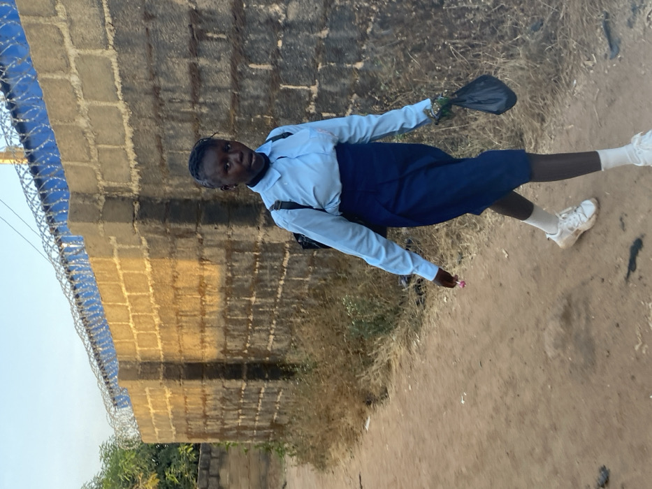 An individual walks along an unpaved dirt path in Kirana, Nigeria. The subject, a young person with dark skin and braided hair, wears a light blue long-sleeved collared shirt, a navy blue high-waisted skirt, white socks, and white athletic shoes. A dark backpack is visible on their back. The individual holds a pink lollipop in their left hand and a crumpled black plastic bag in their right hand. They are looking directly forward, towards the camera. To the right of the path stands a tall wall constructed from grey concrete blocks, partially covered with blue material and topped with razor wire. Sunlight illuminates the upper portion of the wall. Dry, sparse vegetation and some ground debris are present along the path and at the base of the wall. A small amount of green foliage is visible in the upper left background against a clear, light blue sky.