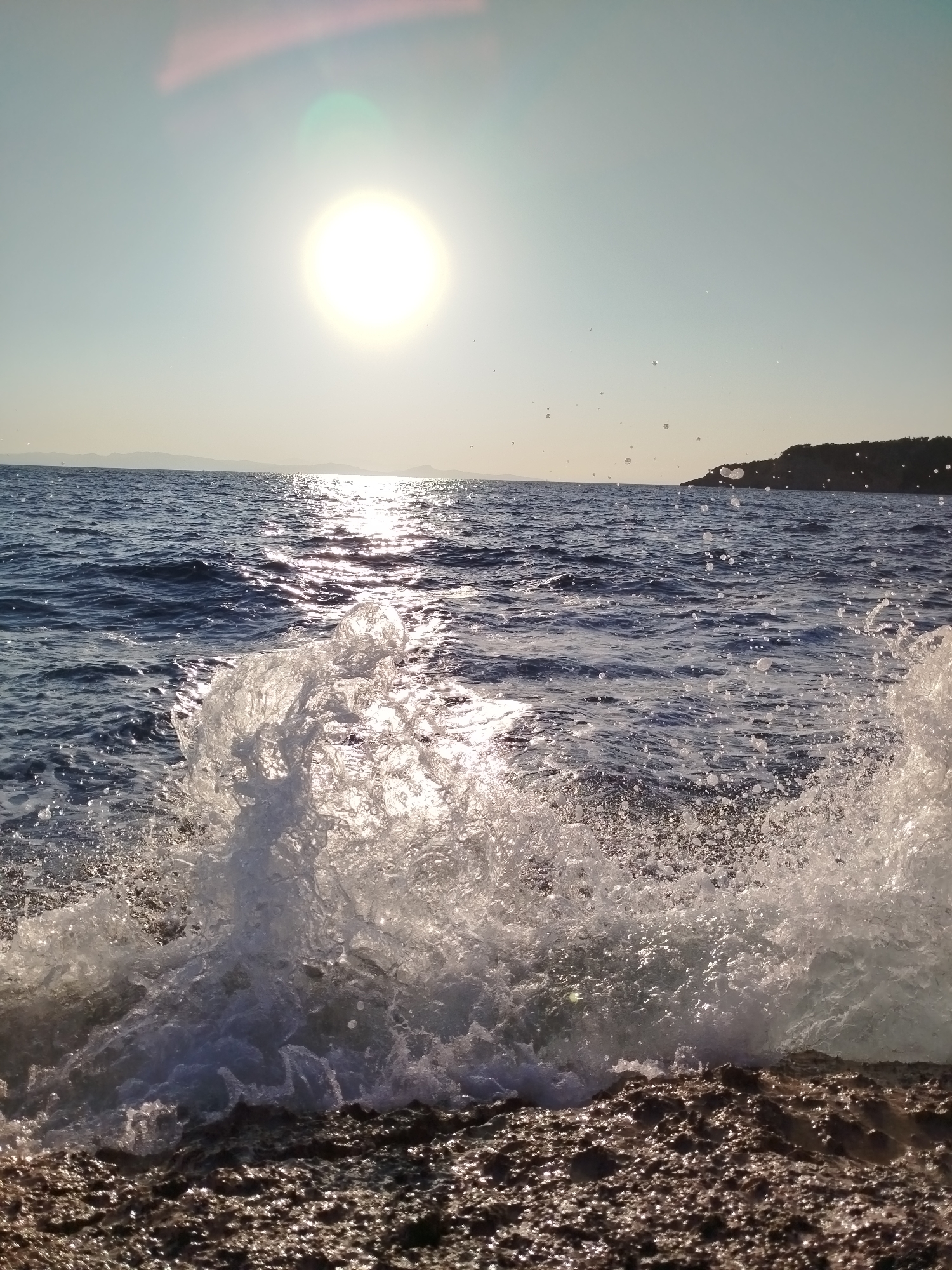 A vertical, eye-level shot captures a wave crashing onto a rocky shore near Karaburun, Turkey, under a bright, hazy sun. The sun, positioned in the upper left quadrant of the frame, is a brilliant, blown-out circle casting a strong glare and reflections across the choppy, dark blue ocean. The water is textured with small waves and scattered white foam. In the foreground, a large wave is breaking, sending white spray and droplets into the air. The shore is comprised of dark, rough rocks, some of which are wet from the spray. In the distance, a dark, tree-covered landmass is visible on the horizon. The sky is a pale, almost white blue, with a slight pinkish hue near the sun due to lens flare. The scene appears to be a natural coastal environment.