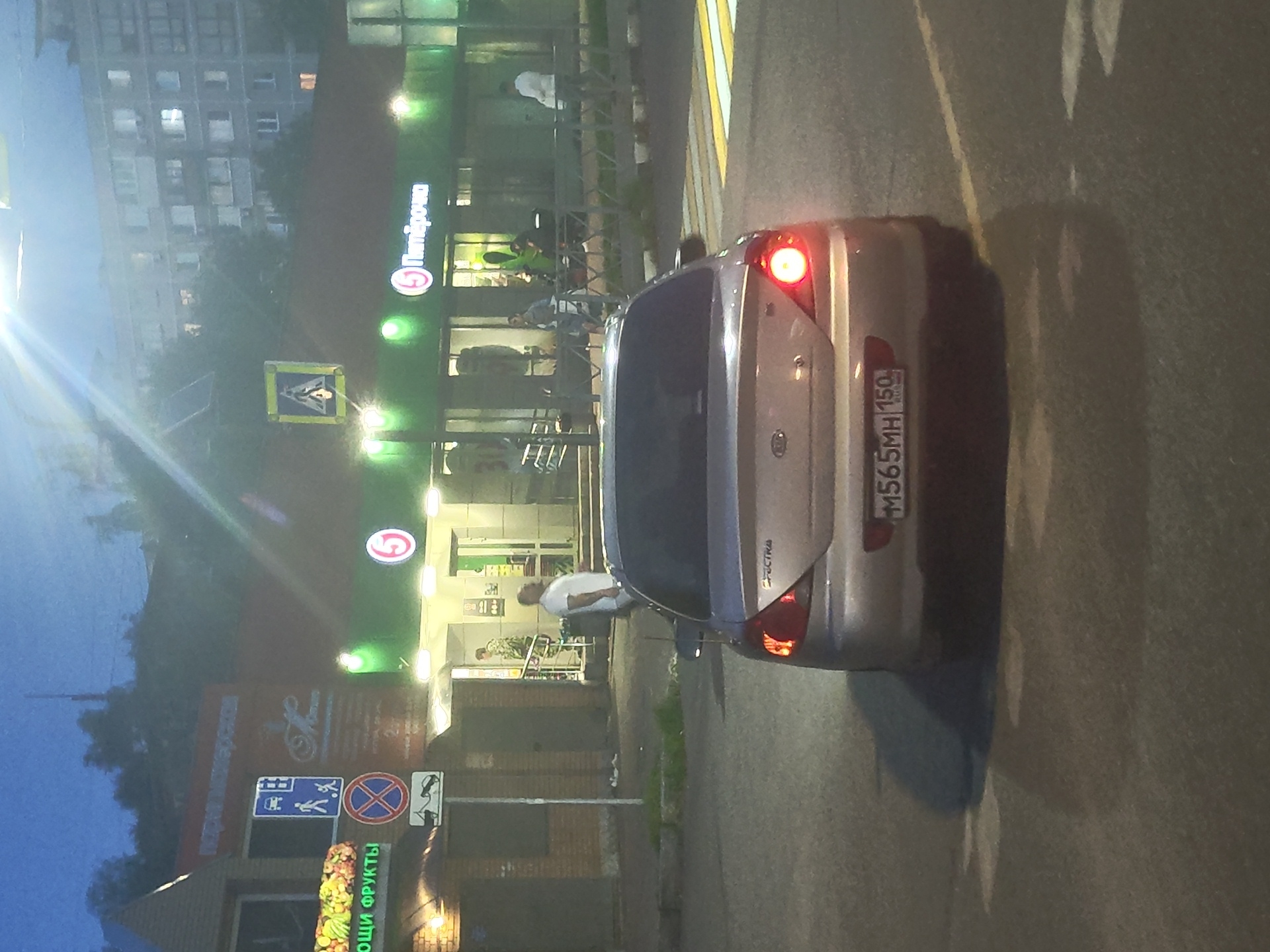 A silver Kia Spectra car is parked at an intersection on a dimly lit street in Ramenskoye, Russia. The car's brake lights are illuminated, indicating it is stopped. In the background, a "Pyaterochka" grocery store is visible with its lights on. Several pedestrians are present near the store entrance. A street sign indicating a pedestrian crossing and a no parking zone are also visible. The overall scene suggests an evening setting in an urban environment.