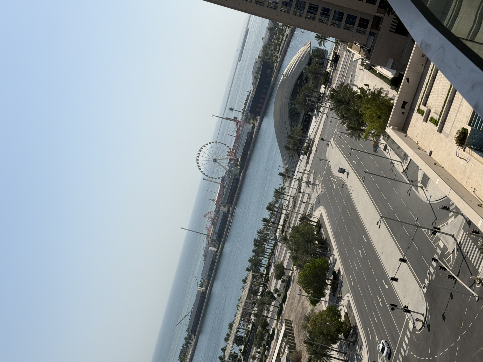 This is a high-angle, full-shot, eye-level, portrait-orientation, outdoor, daylight, medium-level, full-color, long shot photograph taken from a building overlooking a waterfront cityscape. The sky is a pale blue with a slight gradient fading to white. The sea is a calm, light blue, reflecting the sky. In the distance, a large Ferris wheel is visible, along with roller coaster tracks and other amusement park structures. A long, dark building, possibly a pier or a factory, extends into the water.
In the foreground, a multi-lane road curves through the city. White lane markings and arrows are visible. A few traffic lights are positioned at intersections. A single white car is driving on the road. Palm trees and other trees line the sides of the road and are scattered throughout the scene. A modern, curved building with a white roof is situated near the water's edge. To the right, a tall, light-brown building, possibly a hotel or residential tower, dominates the right side of the frame. Part of a balcony railing is visible in the bottom right corner. The overall scene depicts a well-developed urban environment with a significant waterfront attraction.