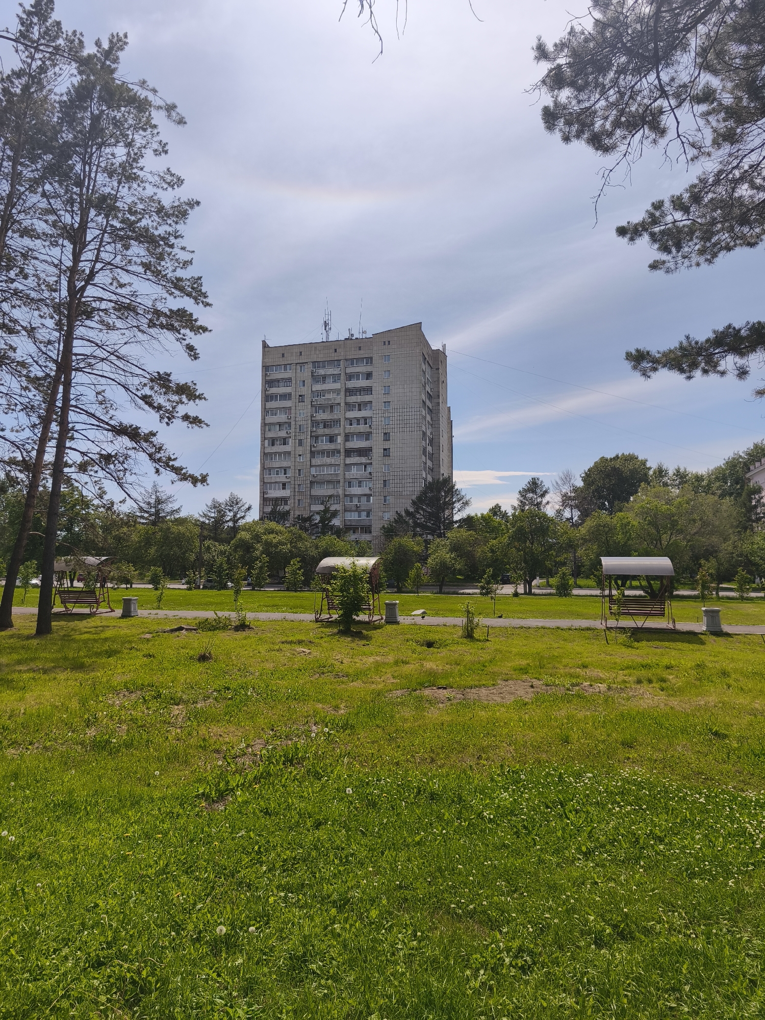 The image depicts an outdoor scene with a tall, light-colored apartment building in the background. The building has many windows and appears to be made of concrete panels. In front of the building, there is a park-like area with green grass and trees. There are two swings with canopies visible in the park. The sky is blue with some scattered clouds, and a faint rainbow is visible. The foreground is dominated by a grassy field with some scattered dandelions. The overall impression is a residential area in a city. Based on visual cues and the context provided, this scene is located in Komsomolsk-on-Amur, Russia.
