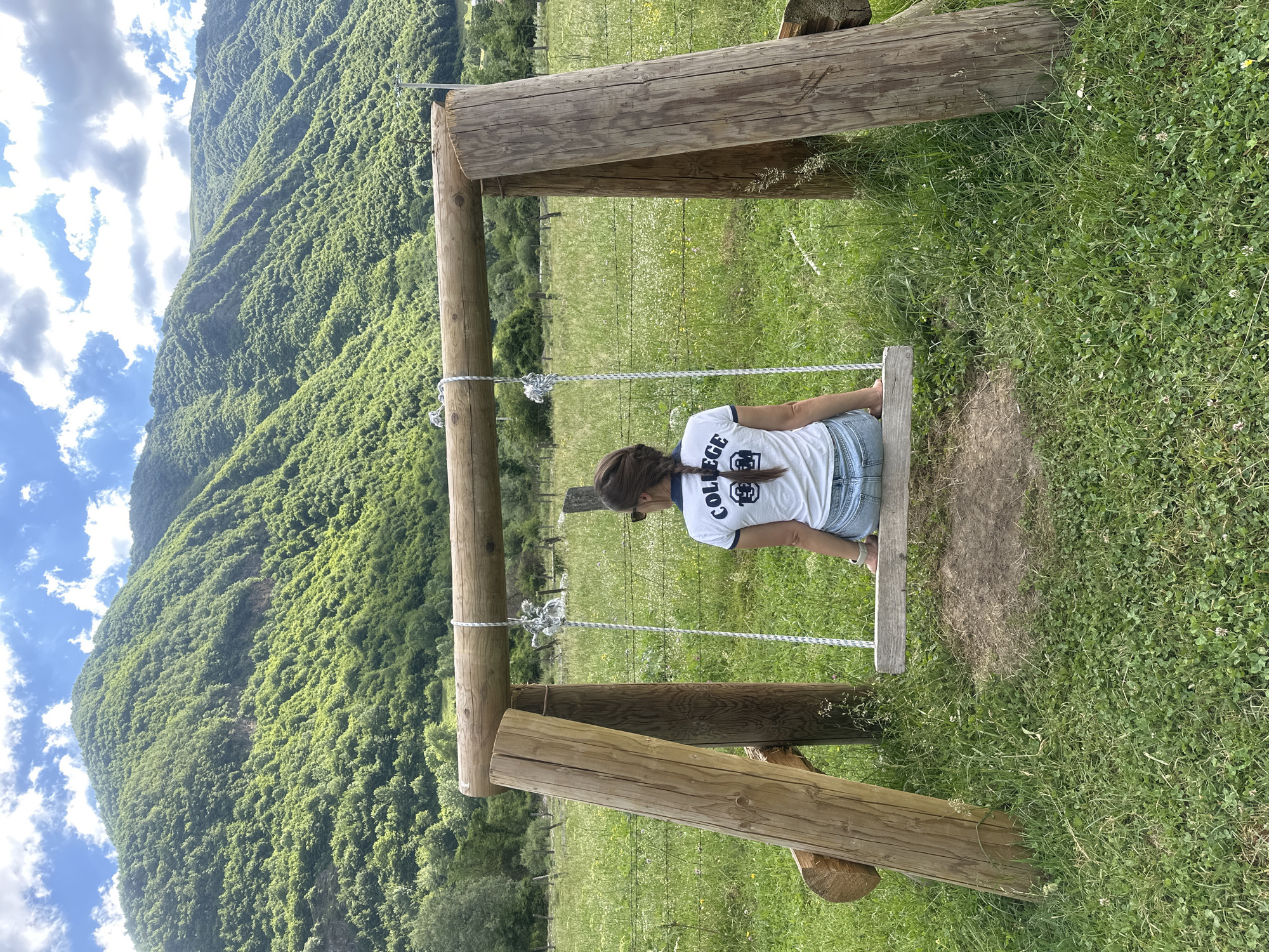 A young woman with dark hair in a braid, wearing a white t-shirt with the word "COLLEGE" printed on it in dark letters and denim shorts, sits facing away from the camera on a wooden swing. The swing is supported by a rustic wooden frame constructed from thick logs. The scene is set in a grassy field with scattered wildflowers. In the background, a dense, green, tree-covered mountain rises under a partly cloudy blue sky. The location is a rural area near Gusinje, Montenegro.