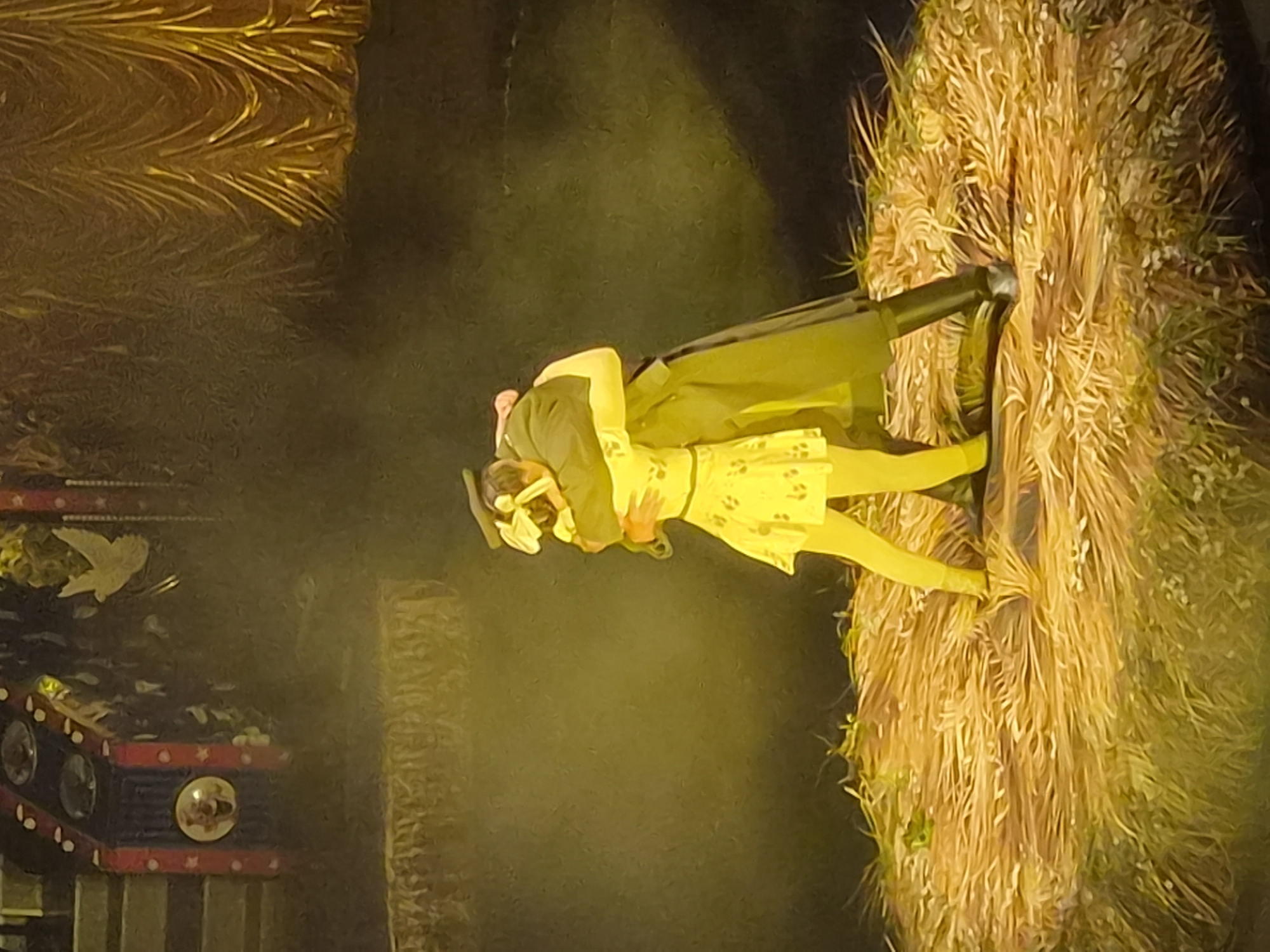Two individuals, one dressed in a dark coat and cap, and the other in a yellow dress and yellow leggings with a yellow bow in their hair, are embracing on a circular platform covered in straw and dried grass. The platform is lit with a warm yellow spotlight, creating a hazy effect in the background. Behind them, to the left, is a stage decoration with red and blue stripes and circular elements, possibly depicting a clown's face. To the right, a golden, draped curtain is visible. The scene appears to be a theatrical performance taking place on a stage. The location is not specified to be in the city of Vladivostok, Russia.