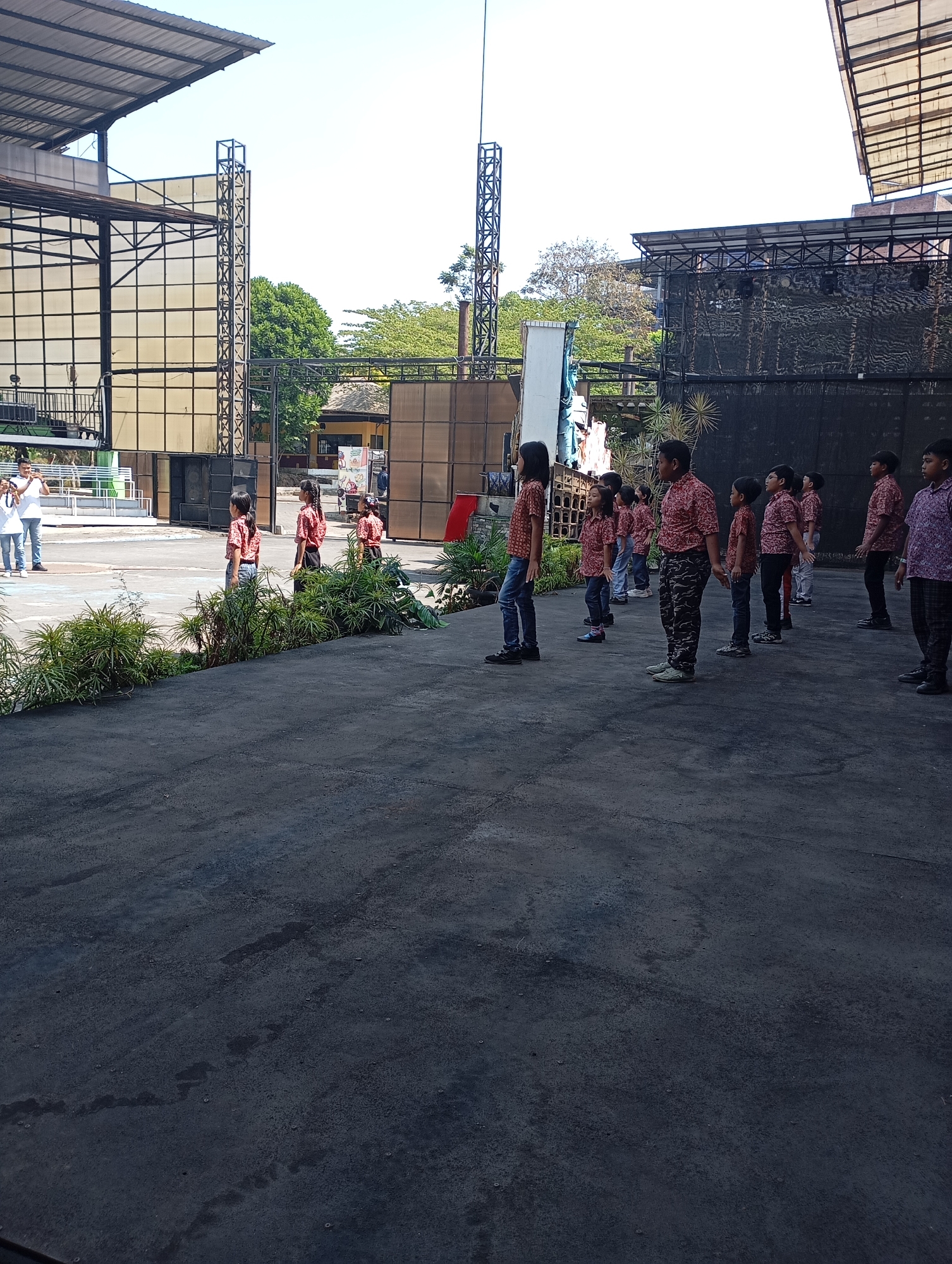 The image captures an outdoor event in Batu, Indonesia, likely at a performance venue. A group of children, mostly boys, are lined up facing away from the camera. They appear to be in uniform, wearing patterned batik shirts. Some are standing attentively, while others seem to be in mid-motion. In the background, a stage structure is visible, along with large speakers and scaffolding. Two adults, a man and a woman, are also present near the stage, with the man appearing to be taking a photo. Lush green plants are placed in the foreground, framing the scene. The ground is paved with asphalt, showing some wear and tear. The sky is clear and blue, suggesting a sunny day.