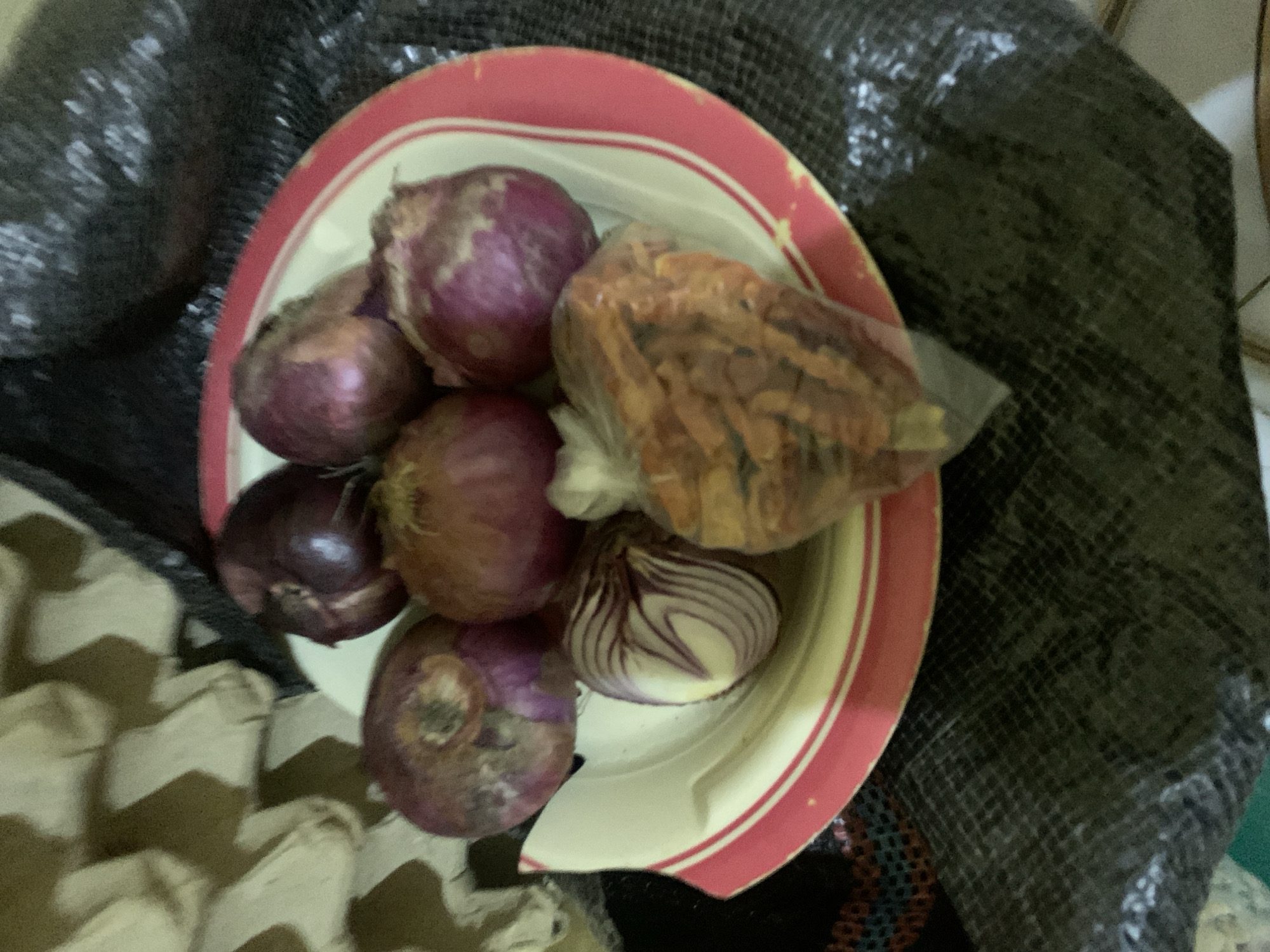The image depicts a red and white plate filled with red onions and a clear plastic bag containing dried chili peppers. The plate is positioned on top of a dark, possibly textured cloth. To the left of the plate is a partially visible egg carton. The objects are situated in Keffi, Nigeria.