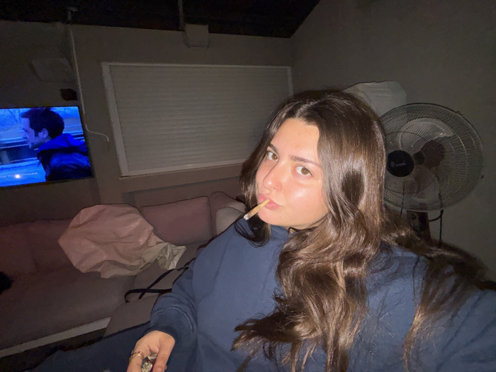 A woman with long brown hair holds a cigarette in her mouth. She wears a navy blue sweatshirt and has rings on her fingers. A television plays in the background, showing a man in a blue jacket. Behind the woman, there is a standing fan and what appears to be a couch with pillows. A white window shade is also visible. The lighting is dim and the setting appears to be indoors.