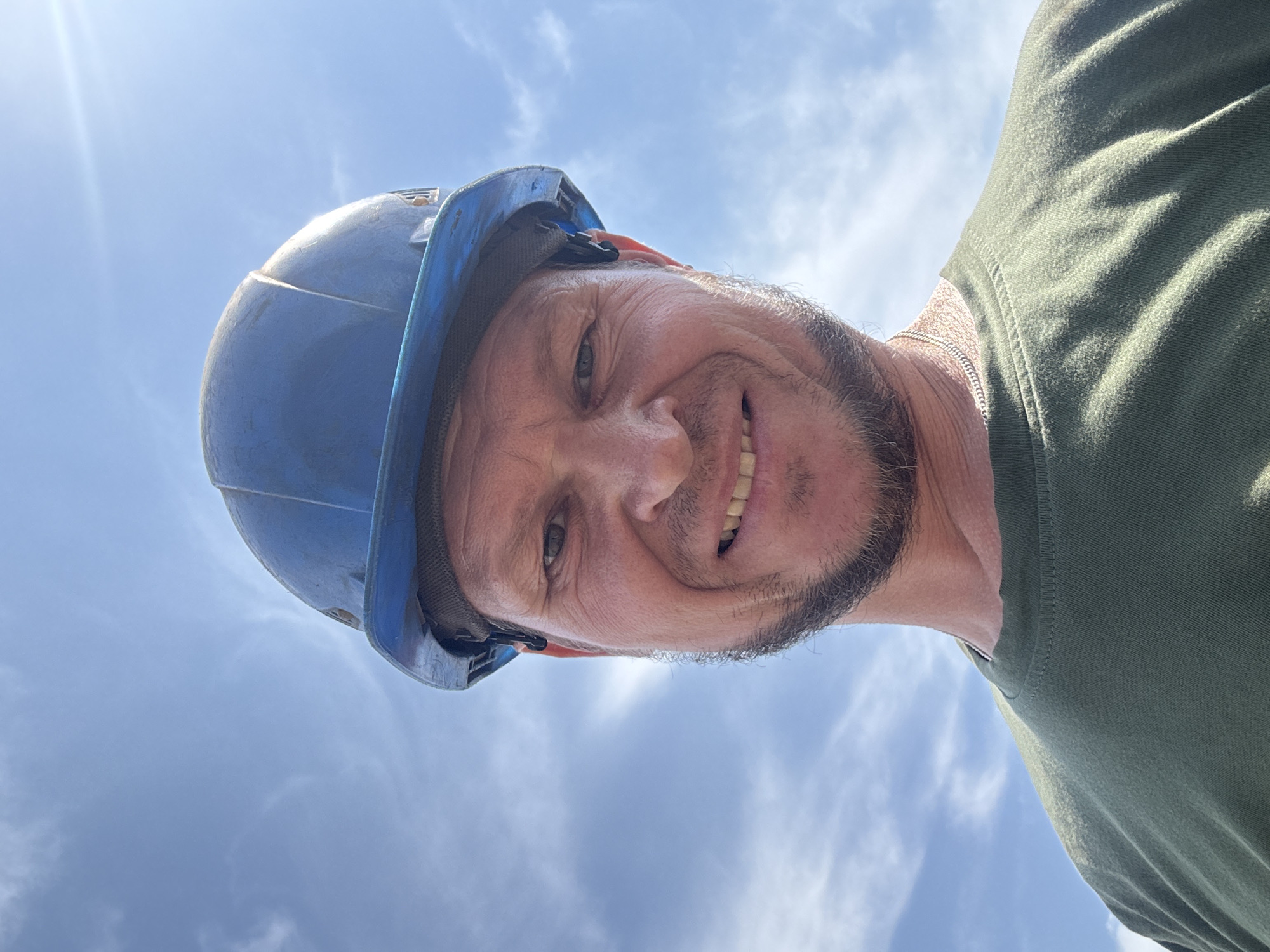 A close-up, low-angle, outdoor selfie shows a Caucasian man from the chest up, wearing a blue hard hat and a green t-shirt. The man has a beard and mustache, and is smiling with his teeth showing. He is wearing a silver chain necklace. The background is a clear blue sky with thin white clouds. The sun is shining brightly, creating a lens flare in the upper right corner of the image. No specific landmarks or features are visible in the background that would indicate a location within Oskemen, Kazakhstan.