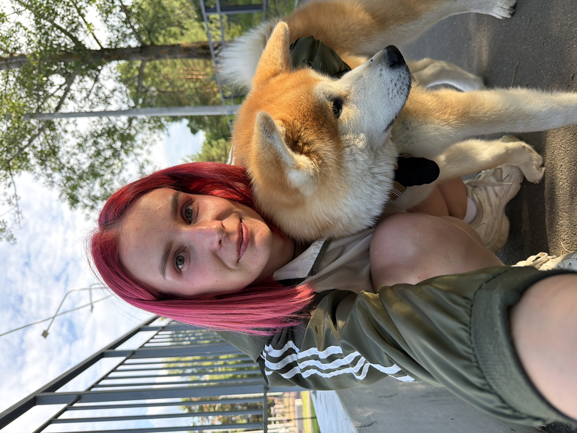 In Chaikovsky, Russia, a young woman with vibrant pink hair is posing for a selfie outdoors. She is wearing a khaki Adidas jacket with white stripes on the sleeves and white sneakers. A large Akita dog, with golden and white fur, is beside her, with its head resting on her arm. The background shows a metal fence, trees, and a cloudy sky, indicating an outdoor setting, possibly a park or a recreational area. The lighting suggests it is daytime.