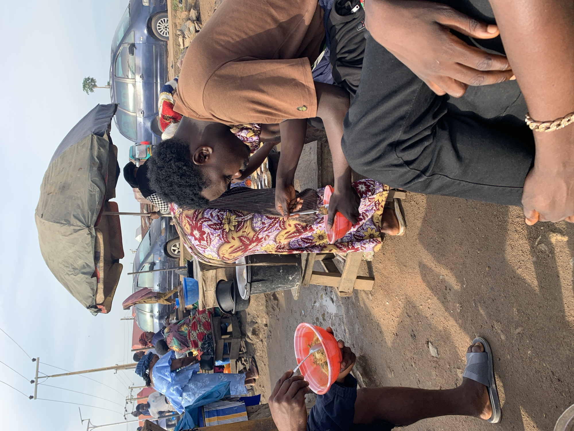 This image depicts an outdoor scene in the city of Rafin Par, Nigeria, likely a market or roadside food stall. In the foreground, a person wearing black pants and a brown t-shirt is seated, holding a red bowl and a spoon, appearing to eat. To their left, another person's legs and feet, wearing gray sliders, are visible. Behind them, a woman in a colorful dress stands, also holding a red bowl and a spoon, suggesting she is eating or serving food. Further back, other individuals are seen in the background, some engaged in selling or preparing food. Several stalls and large black pots are present, indicating a food vending area. A large dark umbrella provides shade. A blue van is parked in the background, along with power lines and utility poles, typical of urban or semi-urban settings. The ground is unpaved, with loose dirt and some debris. The sky is overcast.