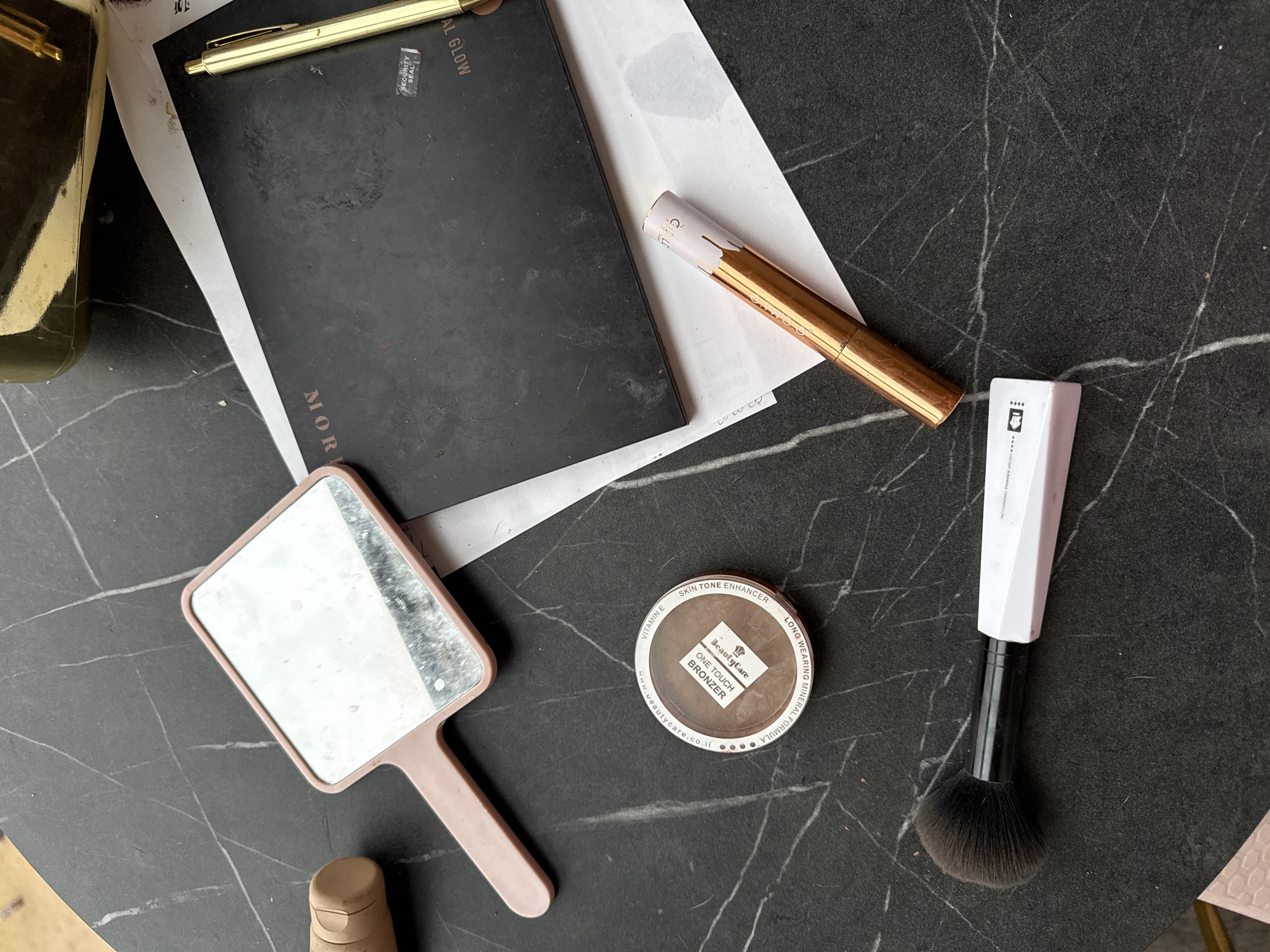 An overhead view captures a collection of cosmetic items and writing implements arranged on a dark, marble-patterned surface in Berezyna, Ukraine. A small, rectangular handheld mirror with a pink plastic frame is positioned centrally on the left, reflecting a distorted view of the surroundings. Below the mirror, a round bronzer compact, labeled "Beauty Care ONE TOUCH BRONZER" with "LONG WEARING MINERAL FORMULA" and "SKIN TONE ENHANCER VITAMIN E" visible on its clear lid, rests on the surface. To the lower left, a makeup brush with black bristles, a black ferrule, and a white, angular handle, partially labeled "POWDER BRUSH," is present.
In the upper right quadrant, a gold-toned retractable ballpoint pen lies horizontally. Below it, a rose gold/copper cosmetic tube with a light purple cap featuring a white "drip" design and the number "232" is positioned diagonally. Partially obscured by the pen and the cosmetic tube is a rectangular black object, likely a makeup palette or a book, displaying fragmented text "MORP" and "IAL GLOW," along with a "SECURITY SEAL" sticker. White papers are visible underneath the black object and the rose gold tube. The dark surface exhibits numerous white, irregular veins, characteristic of a marble or granite pattern. No individuals are present in the scene.