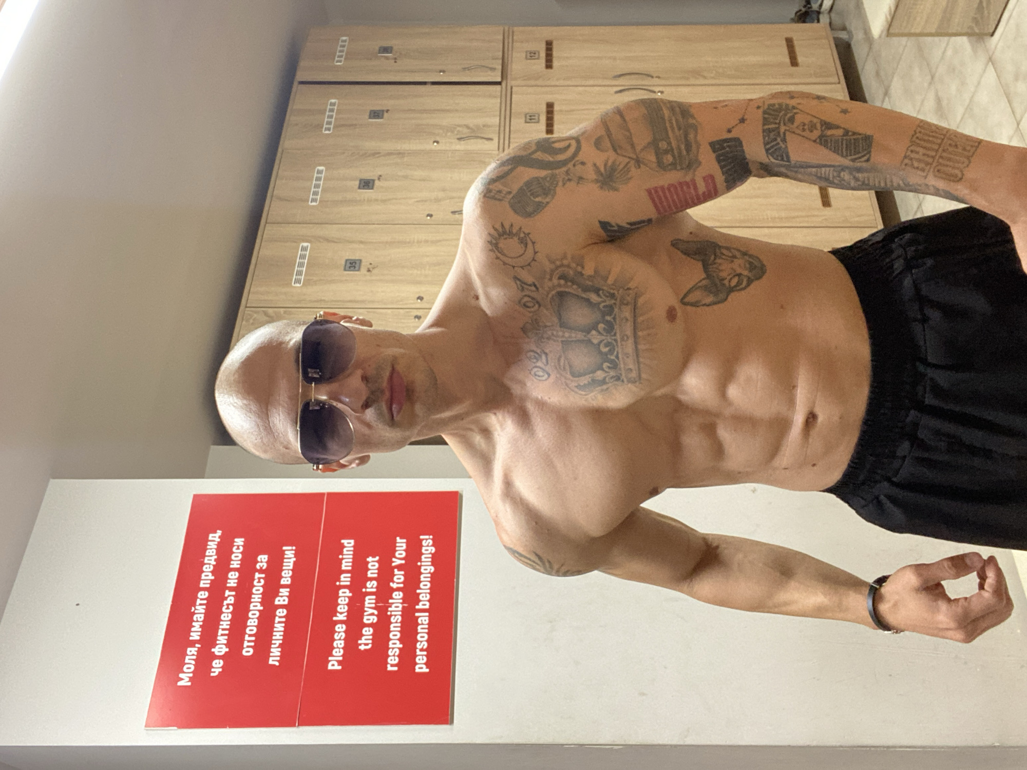 A bald, muscular man with sunglasses and numerous tattoos poses shirtless in front of lockers and a red sign in a gym. The sign is written in both Bulgarian and English, stating that the gym is not responsible for personal belongings. The location is likely Pleven, Bulgaria, as the Bulgarian text on the sign indicates.